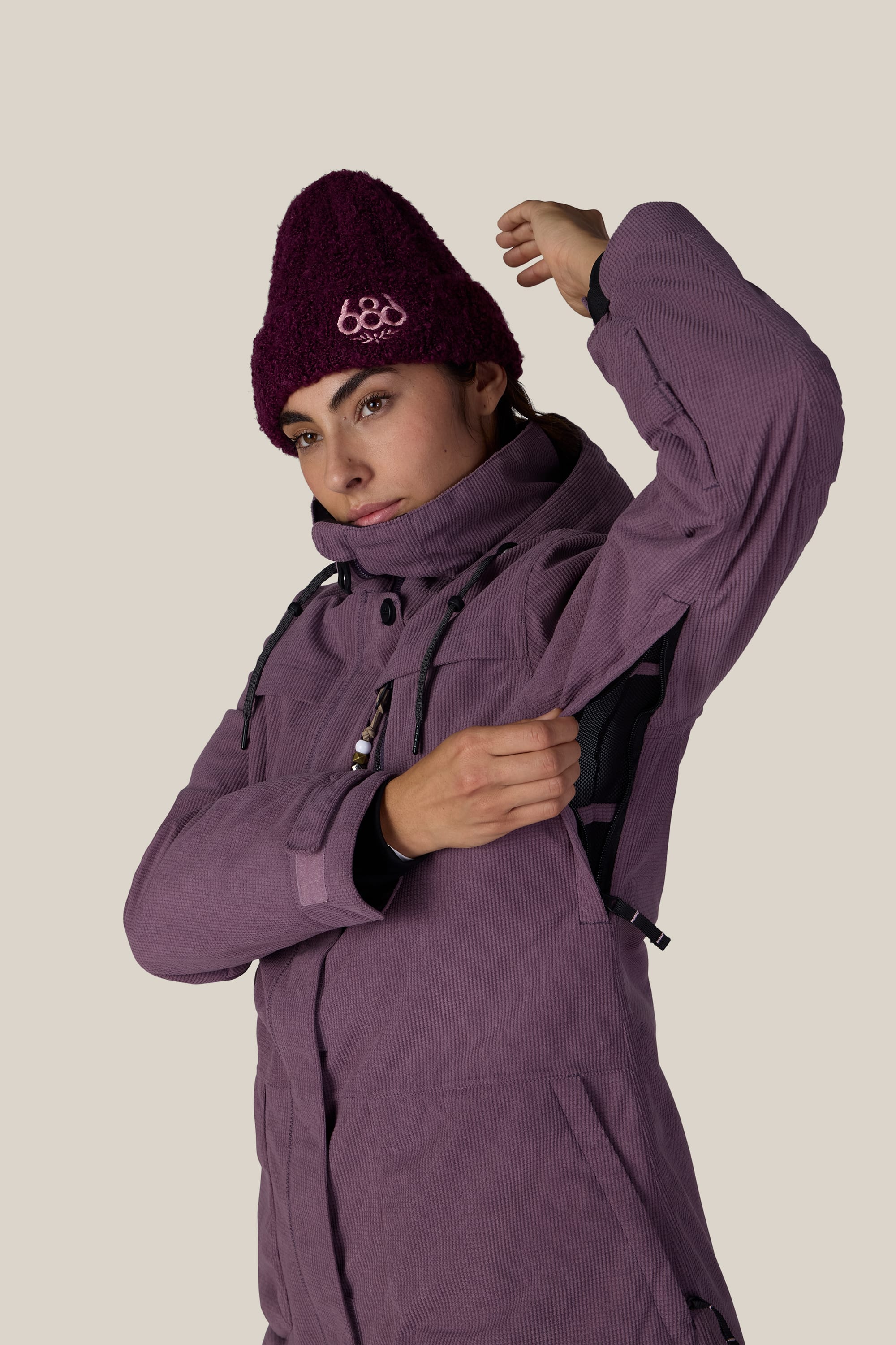 Wearing the 686 Women’s Spirit Insulated Jacket, a person in a maroon knit hat unzips the underarm vent of their purple 686 jacket while standing against a plain beige background.