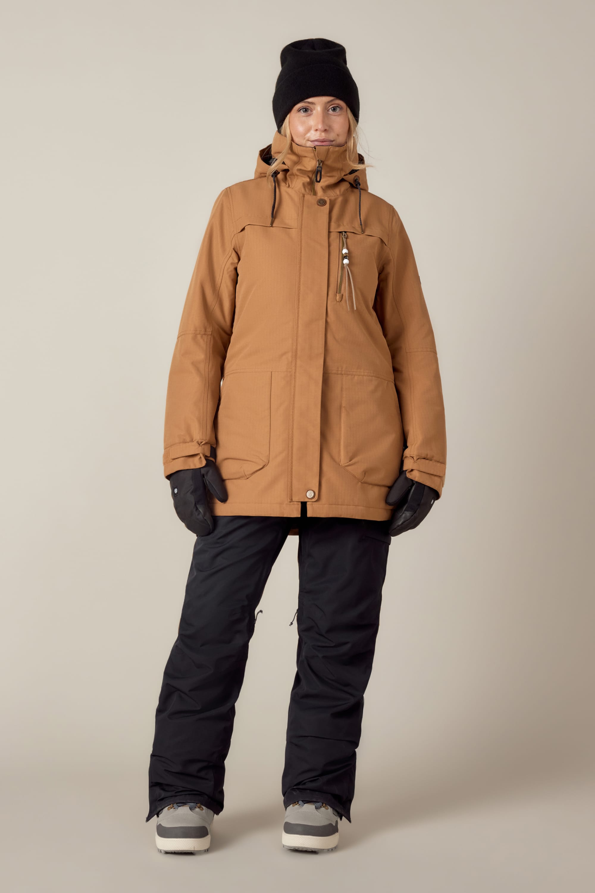 A person in the 686 Womens Spirit Insulated Jacket by 686, paired with black gloves, snow pants, gray boots, and a black beanie, stands against a plain light background.