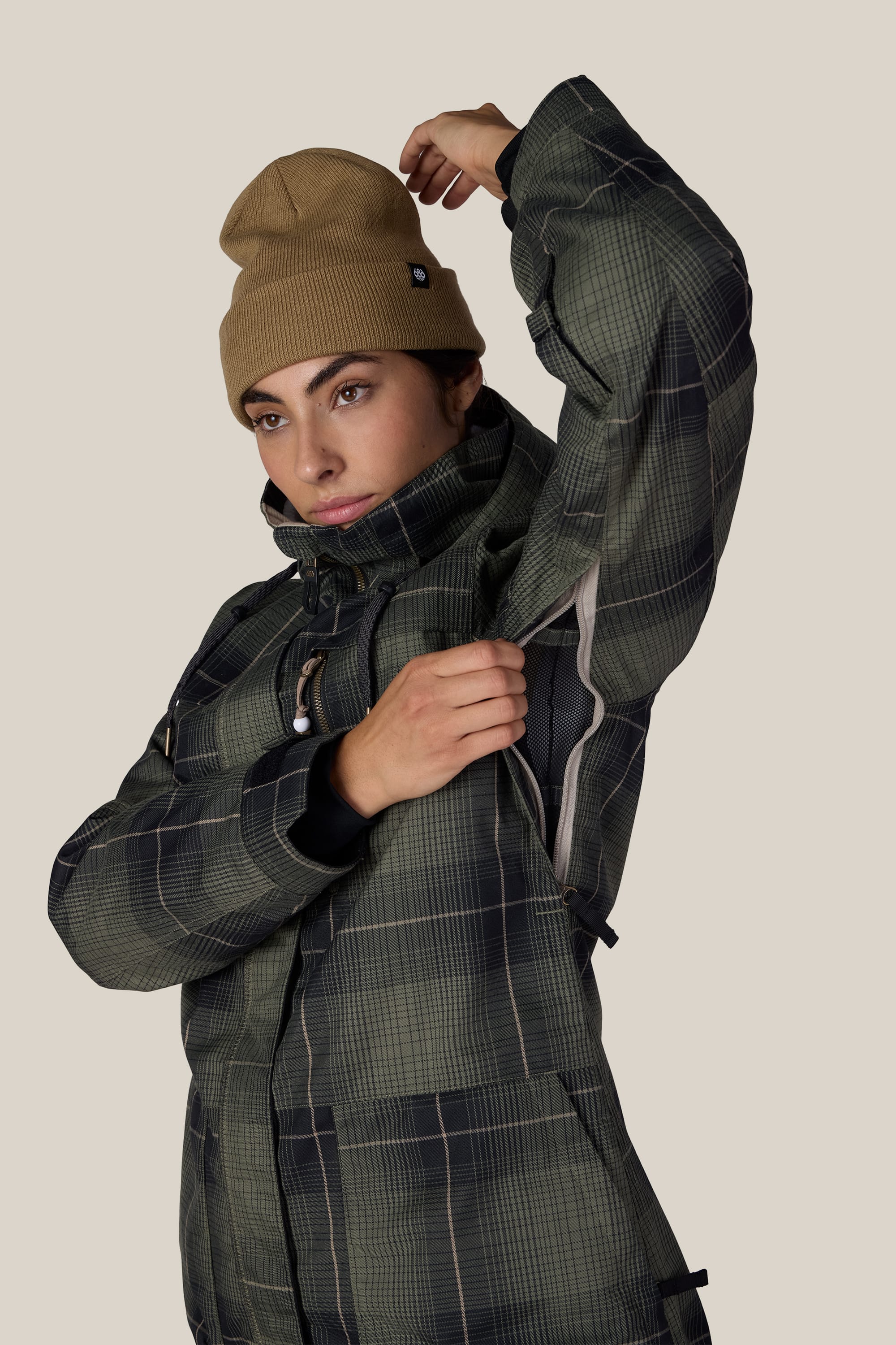 A person wearing the 686 Women’s Spirit Insulated Jacket in green plaid and a tan beanie unzips an underarm vent. The background is plain beige.