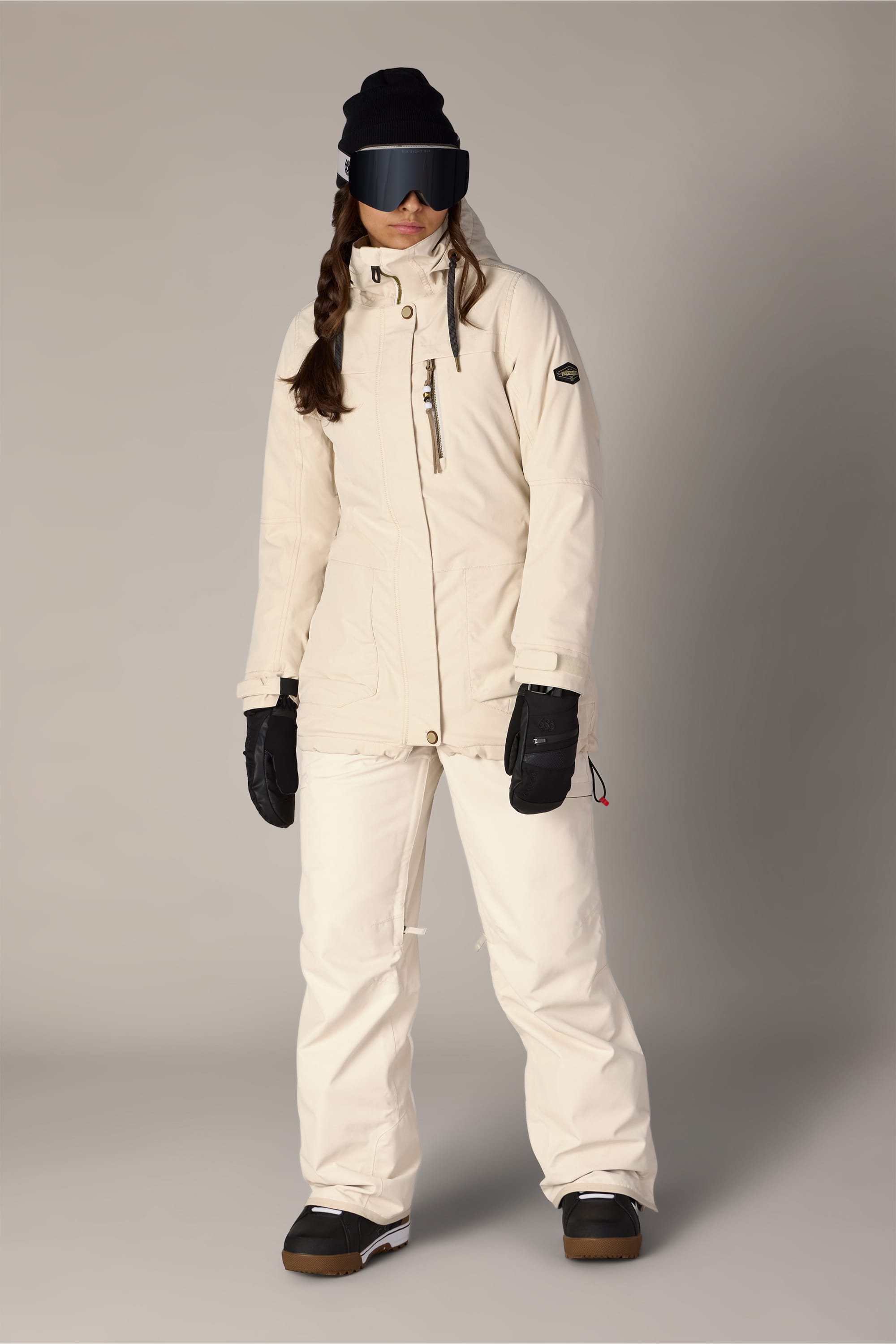 A person wearing the 686 Women’s Spirit Insulated Jacket in cream, paired with matching pants, black gloves, black winter boots, a black beanie, and large ski goggles stands against a plain light-grey background.