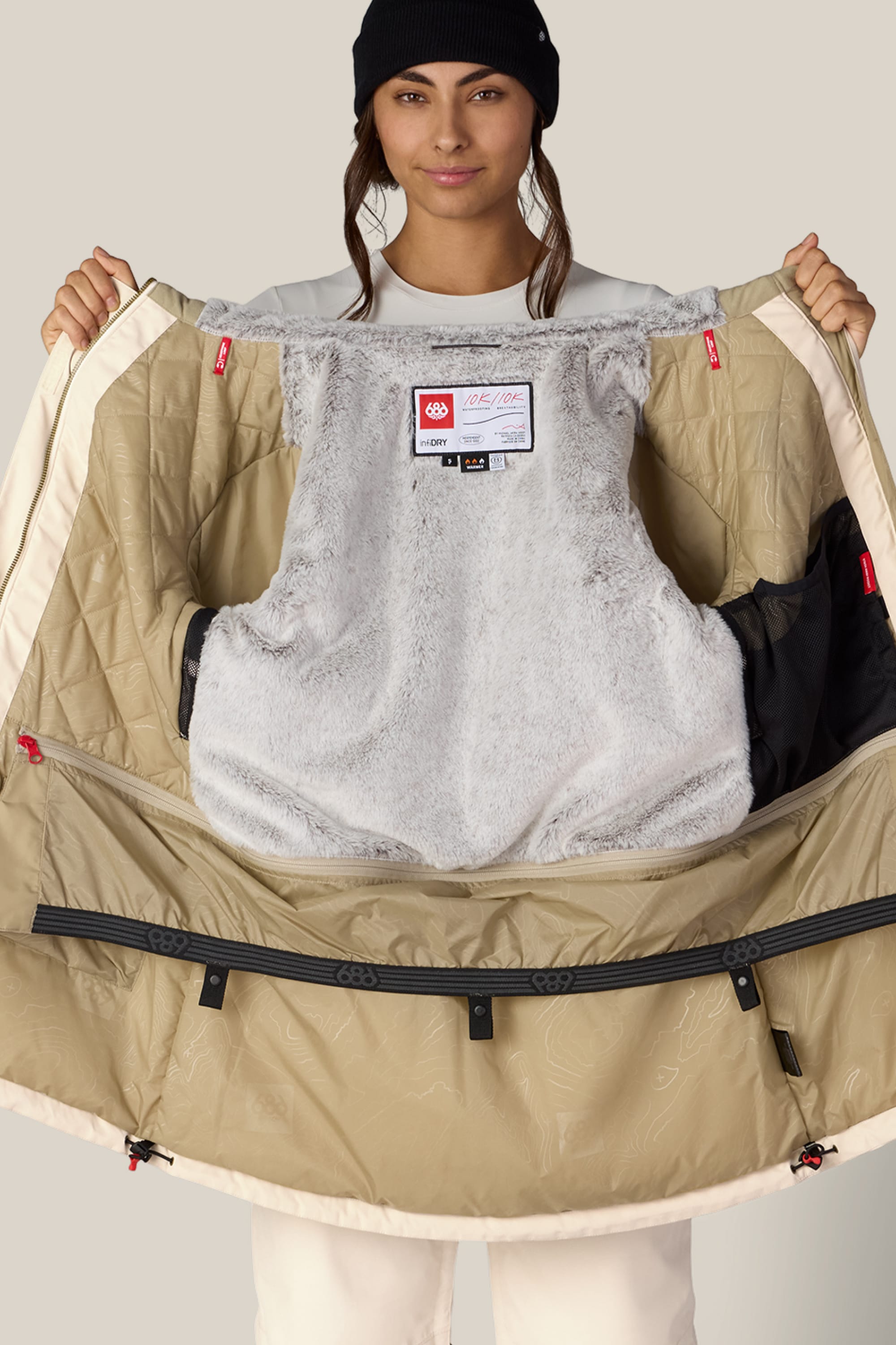 A person wearing a black beanie holds open the 686 Women’s Spirit Insulated Jacket by 686, revealing its light gray furry lining and inner pockets. The person faces forward with a slight smile.