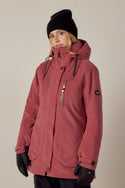 A person wearing the 686 Womens Spirit Insulated Jacket by 686 stands against a plain light background, looking slightly to the side and smiling subtly. They pair it with a black beanie and gloves for a cozy, stylish winter look.