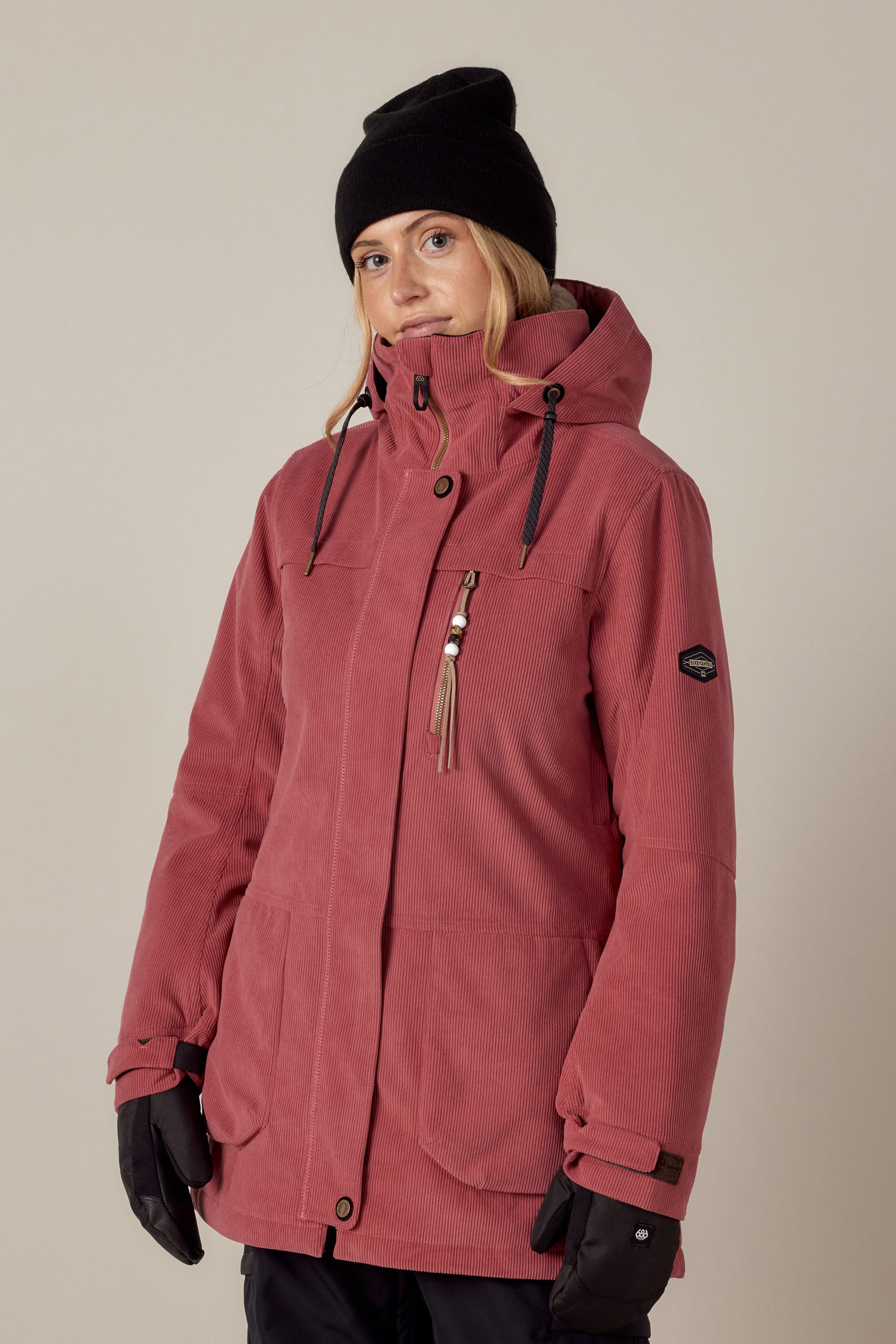 A person wearing the 686 Womens Spirit Insulated Jacket by 686 stands against a plain light background, looking slightly to the side and smiling subtly. They pair it with a black beanie and gloves for a cozy, stylish winter look.