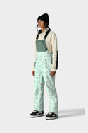 A person in a black beanie, cream hoodie, and 686 x Hello Kitty Womens Harper Shell Bib sage green overalls with pastel splatter patterns by 686 and black Vans sneakers stands against a plain light background.