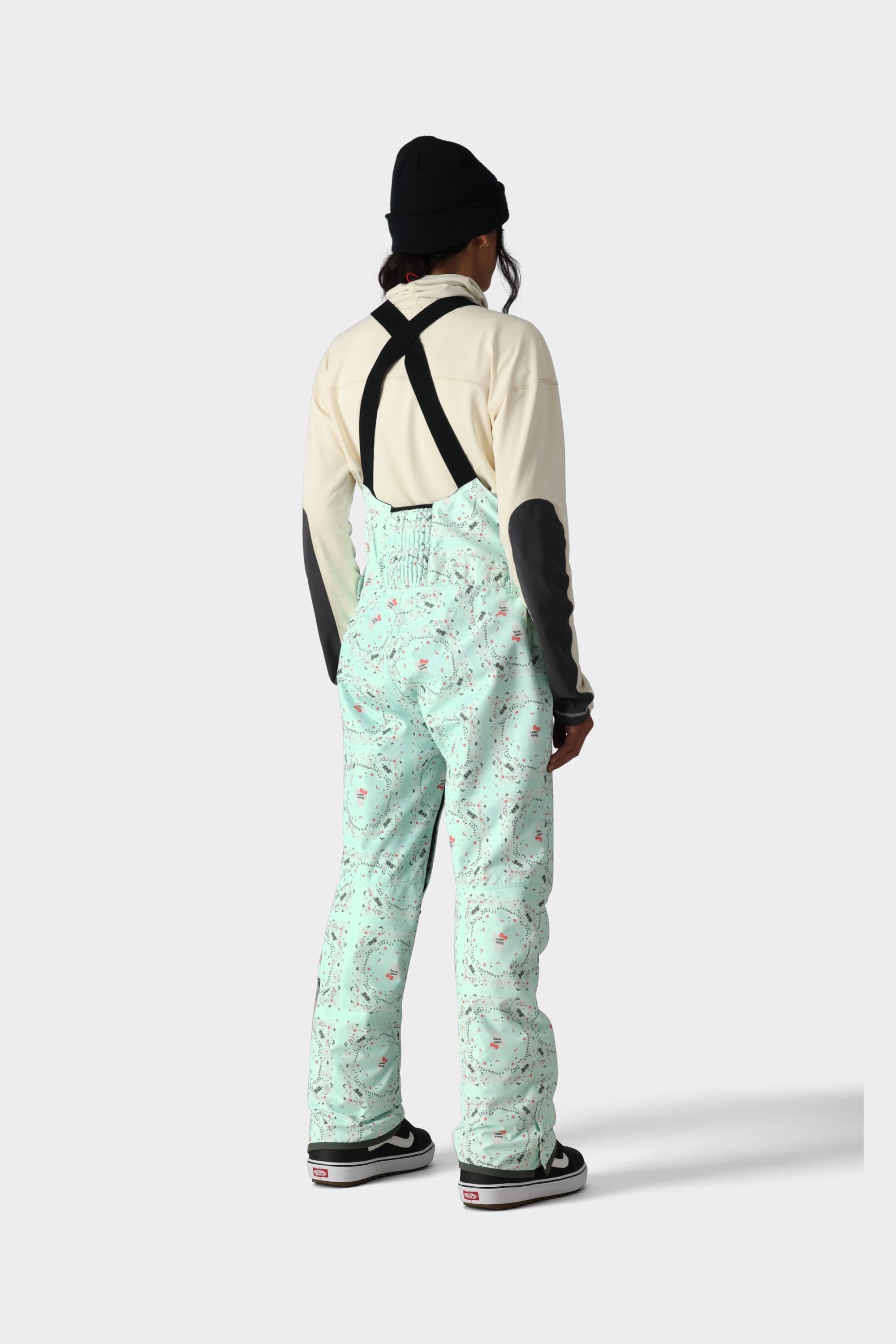 A person faces away, wearing the 686 x Hello Kitty Womens Harper Shell Bib by 686 with black suspenders, paired with a black beanie and a beige jacket featuring black elbow patches. The backdrop is plain light grey.