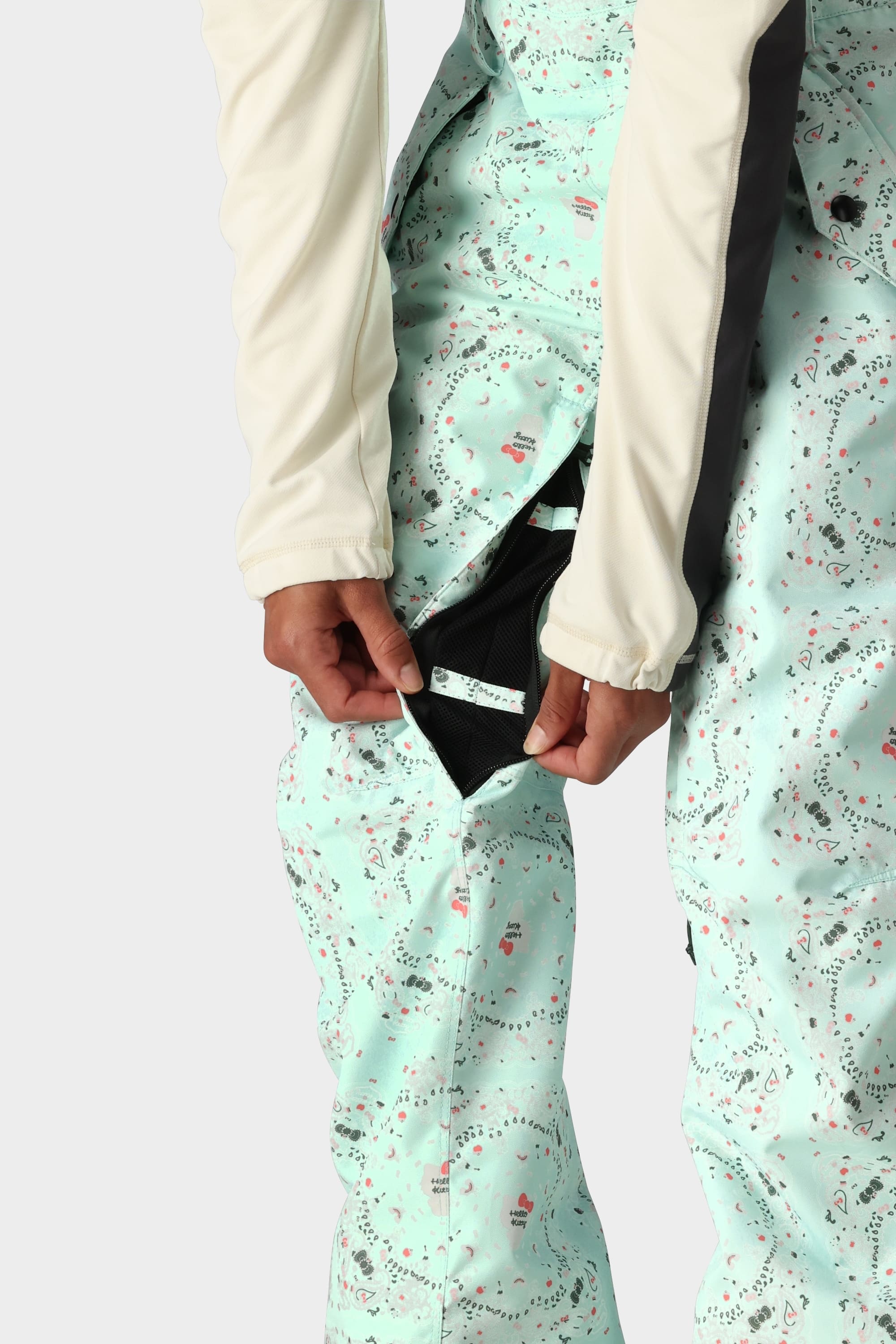 A person in 686 x Hello Kitty Womens Harper Shell Bib, featuring a light mint-green pattern and 10k waterproofing, unzips a side vent to reveal black mesh lining, paired with a cream long-sleeve top.