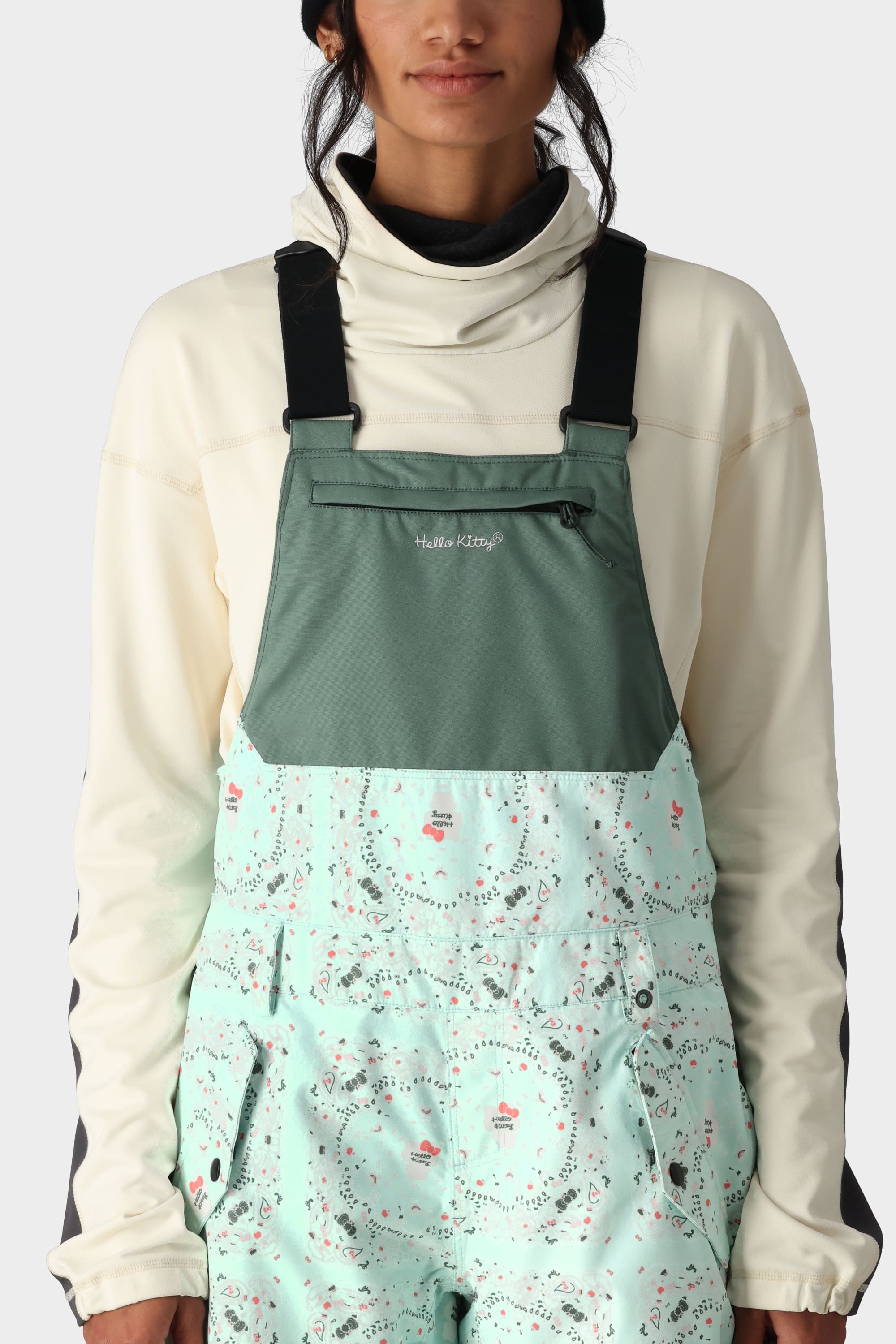 A person wears the 686 x Hello Kitty Womens Harper Shell Bib, a mint green, budget-friendly overall by 686 featuring a playful winter pattern, black straps, a green chest pocket, and Hello Kitty logo over a cream hoodie. Image is shoulder-cropped.