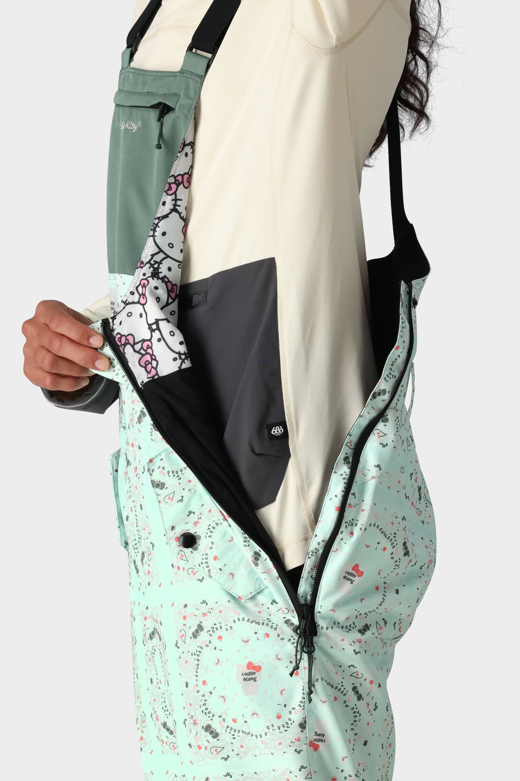 A person wearing the 686 x Hello Kitty Womens Harper Shell Bib by 686 in a cream top unzips a side zipper to reveal a pocket with a Hello Kitty design and grey flap; only their torso and hand are shown.