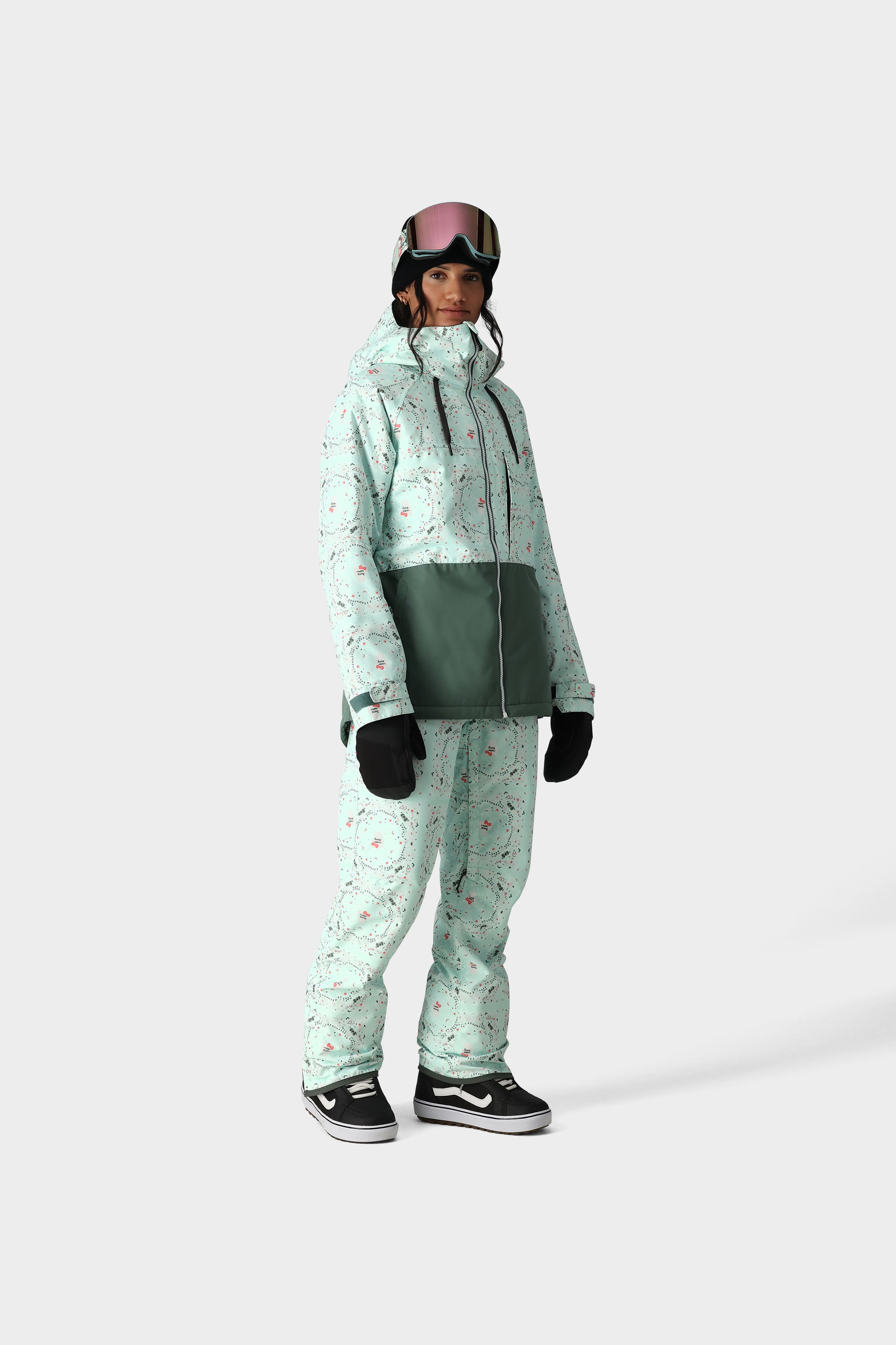 A person stands against a white background wearing the 686 x Hello Kitty Womens Harper Shell Bib by 686, a mint green floral-patterned set with 10k waterproofing, styled with black gloves, black Vans sneakers, and ski goggles—ideal for layering on the slopes.