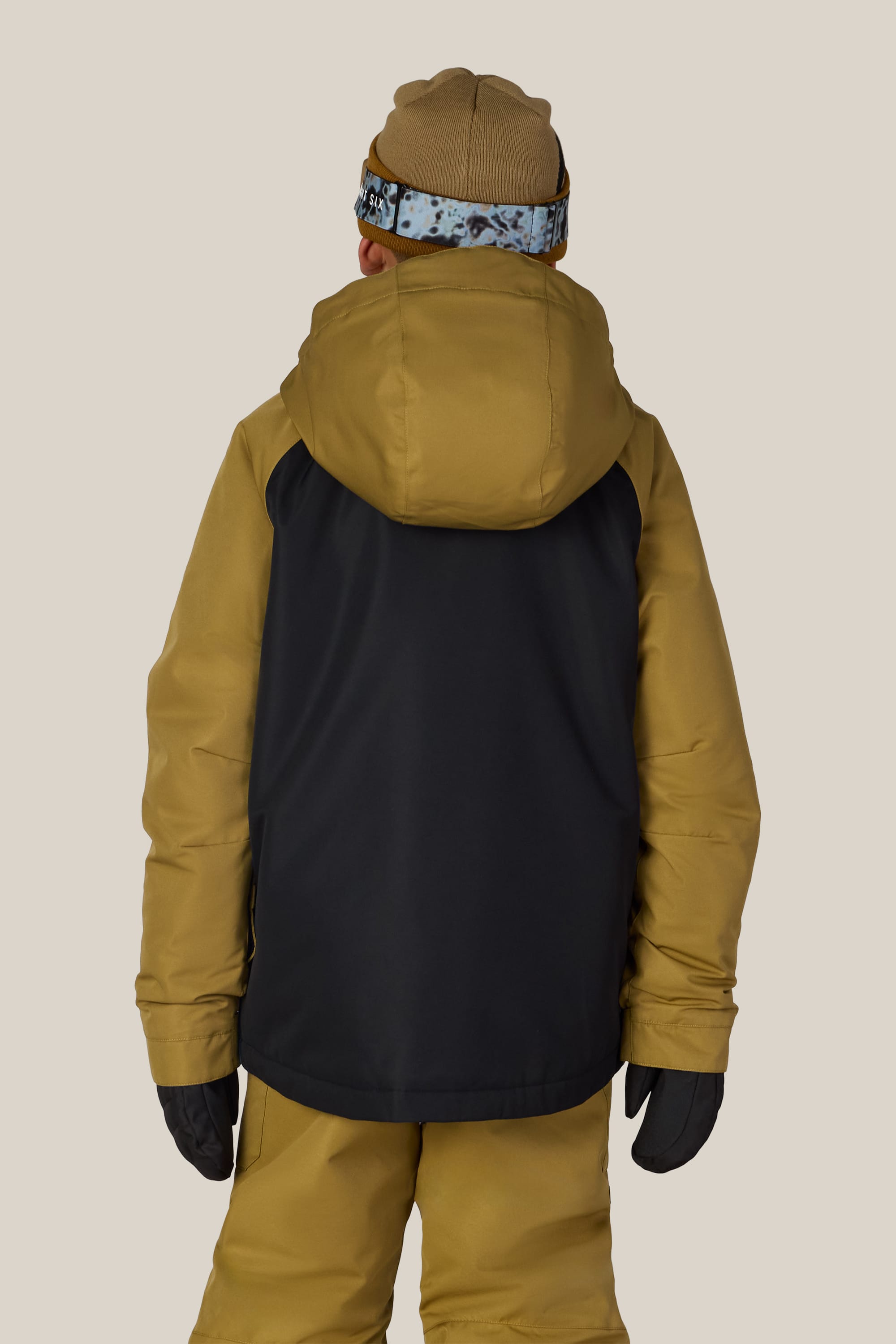 A person wearing the 686 Boys’ Geo™ Insulated Jacket by 686, along with a brown hat, matching pants, and black gloves, is standing with their back to the camera against a beige background.