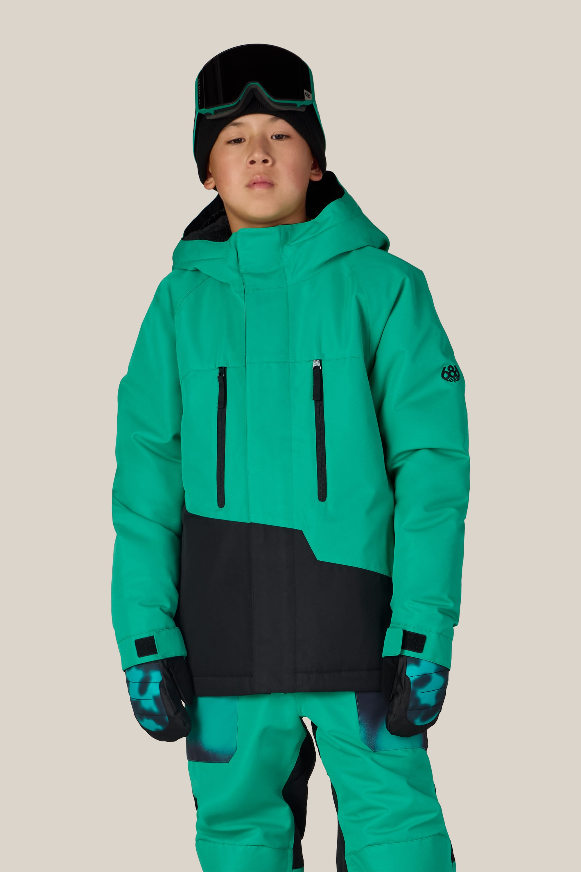 A young person is wearing the 686 Boys’ Geo™ Insulated Jacket by 686 in green and black, paired with matching pants, gloves, and black ski goggles while standing against a plain beige background.