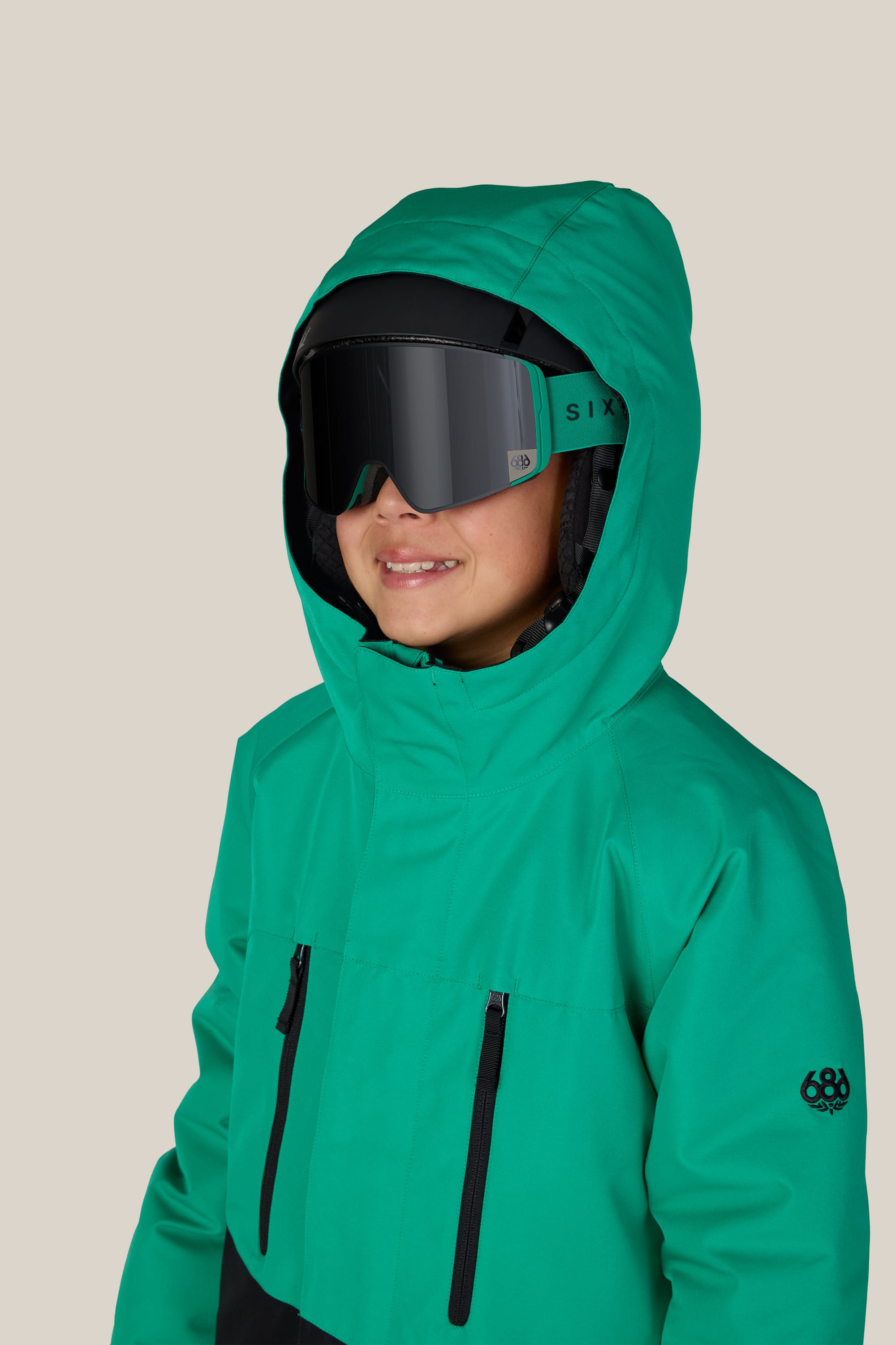 A child dressed in the 686 Boys’ Geo™ Insulated Jacket and large black ski goggles smiles while facing slightly to the right against a plain, light background.