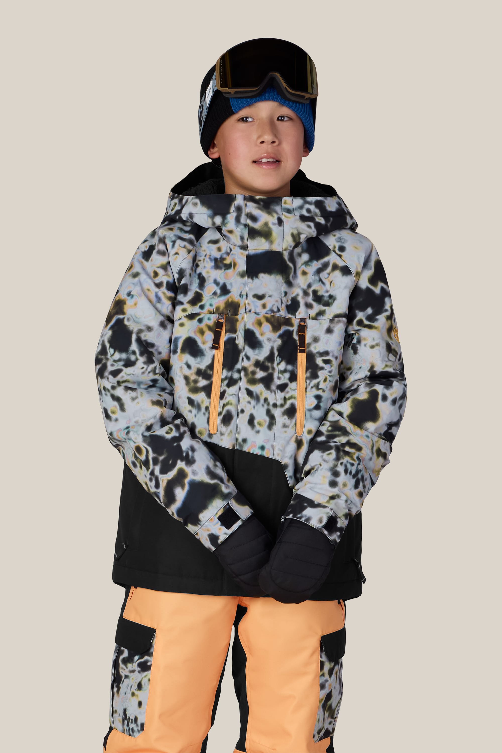 A child wearing the 686 Boys’ Geo™ Insulated Jacket by 686, paired with orange snow pants featuring black patches, black gloves, and ski goggles on their head, stands against a plain light background.