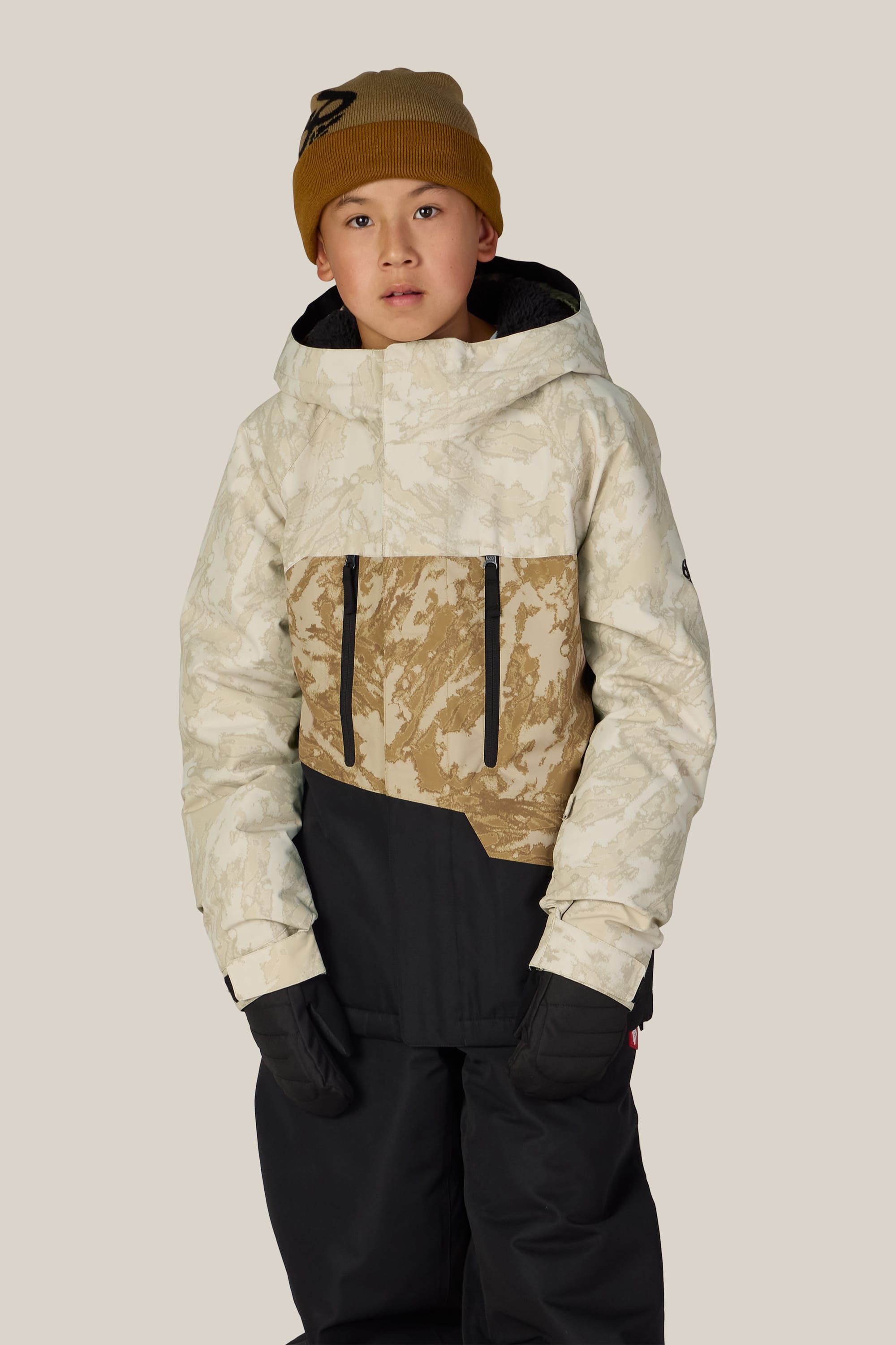 A young person wearing a tan beanie, black gloves, black snow pants, and the 686 Boys’ Geo™ Insulated Jacket by 686 in a beige and tan camouflage pattern stands against a plain light background, looking forward with a neutral expression.