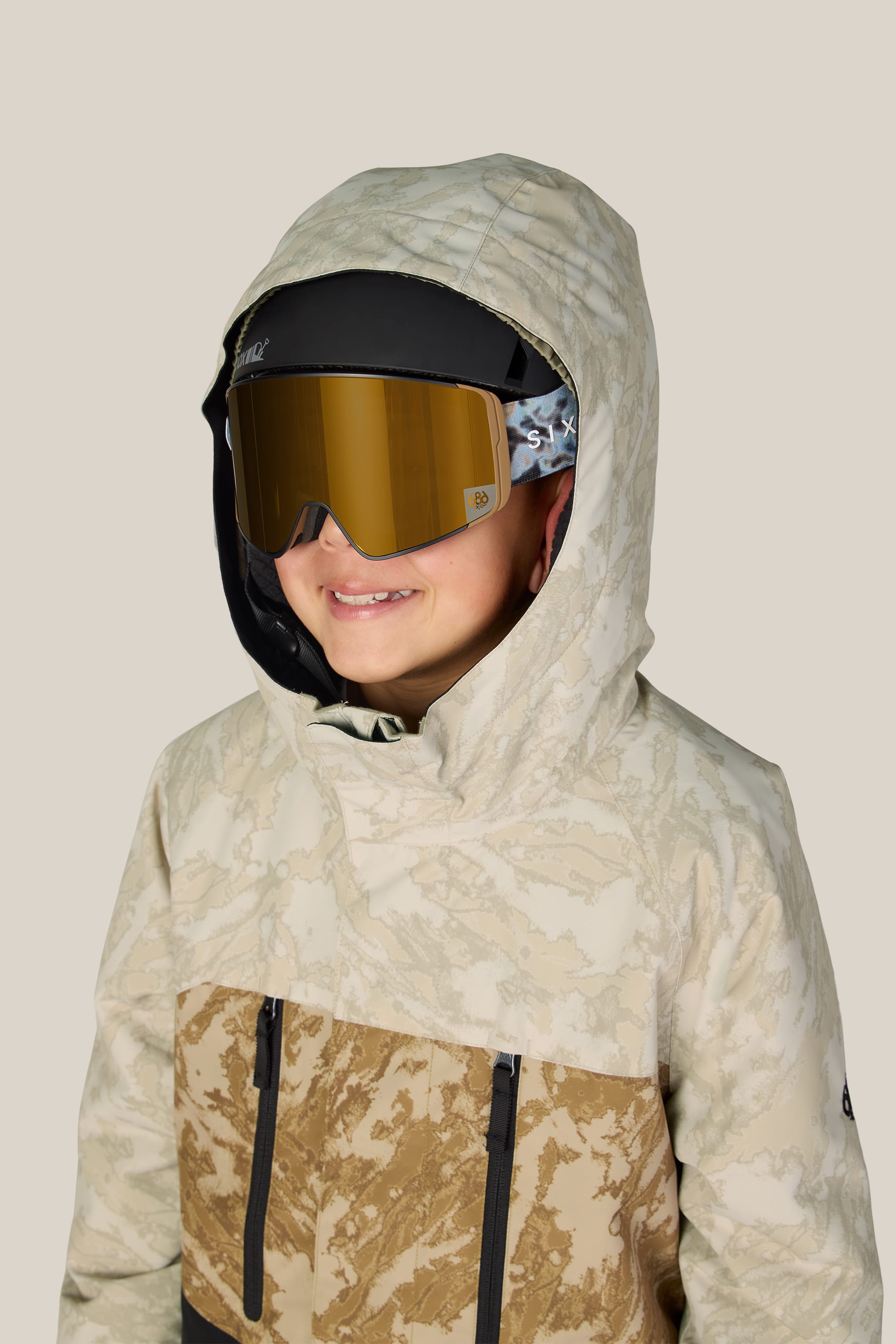 A child smiles against a plain light background, wearing the 686 Boys’ Geo™ Insulated Jacket by 686, paired with dark ski goggles with gold lenses and a black hat.