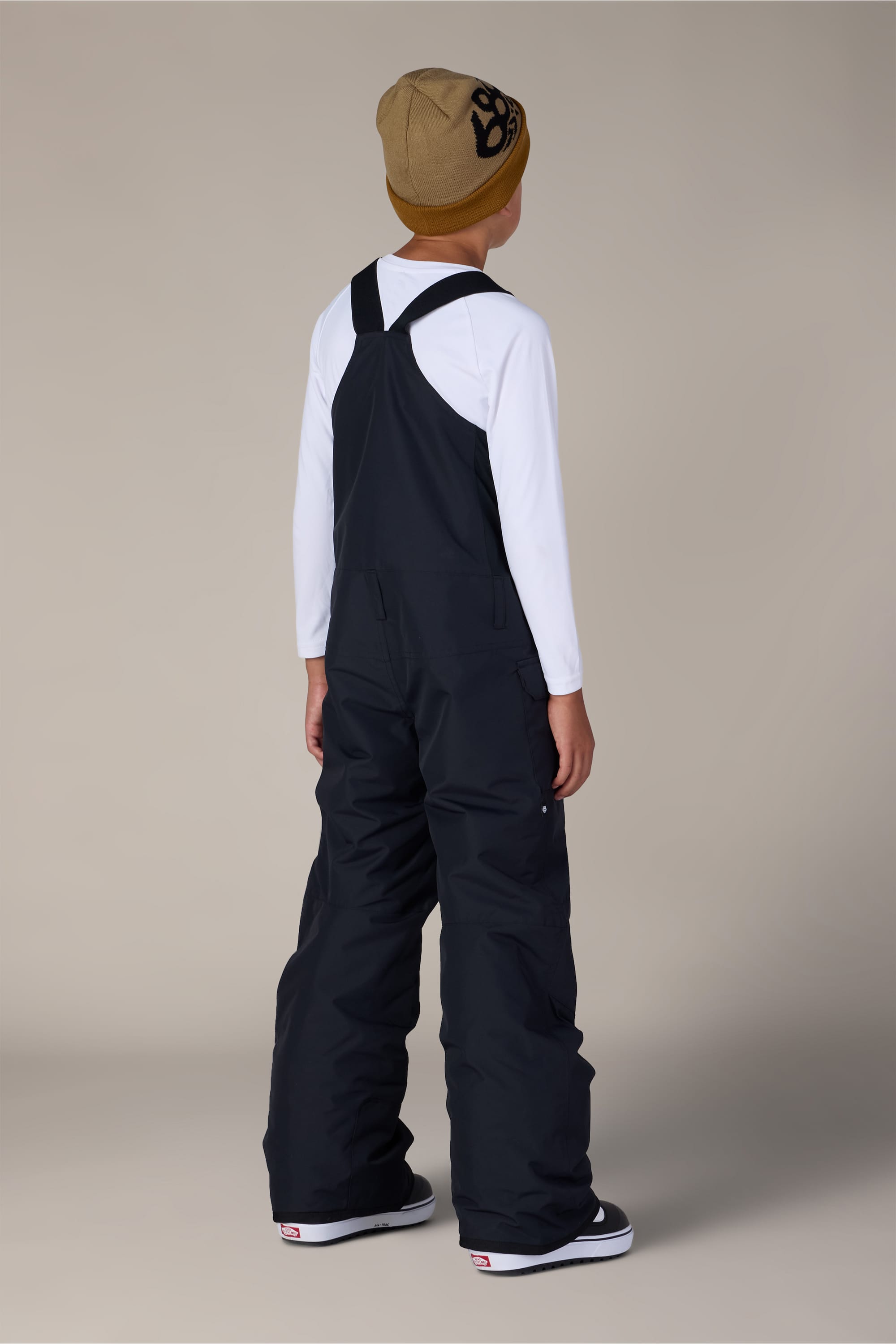 A child wearing a tan beanie, white long-sleeve shirt, and 686 Boys’ Frontier™ Insulated Bib by 686 stands facing away against a plain background.