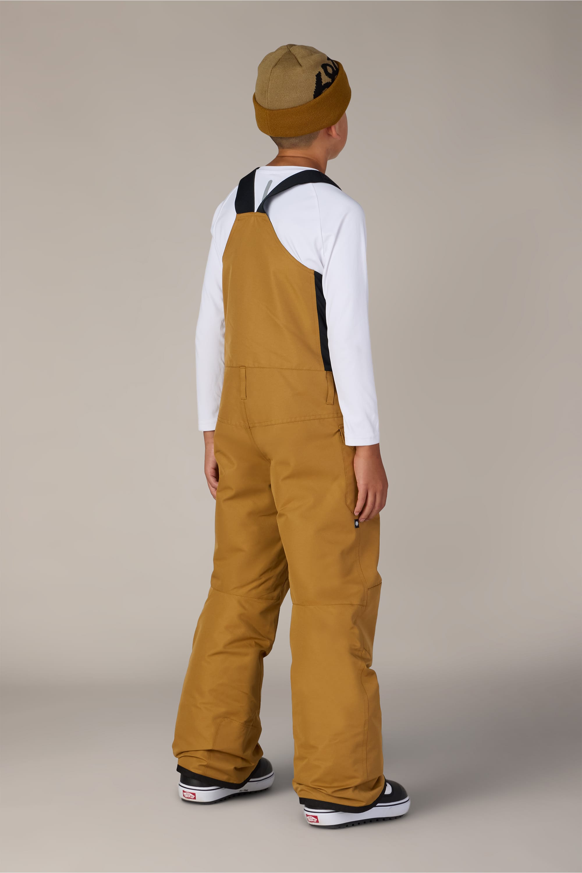 A child stands facing away, wearing the 686 Boys’ Frontier™ Insulated Bib by 686 over a white long-sleeve shirt, paired with a tan beanie and black-and-white sneakers, against a plain light background.