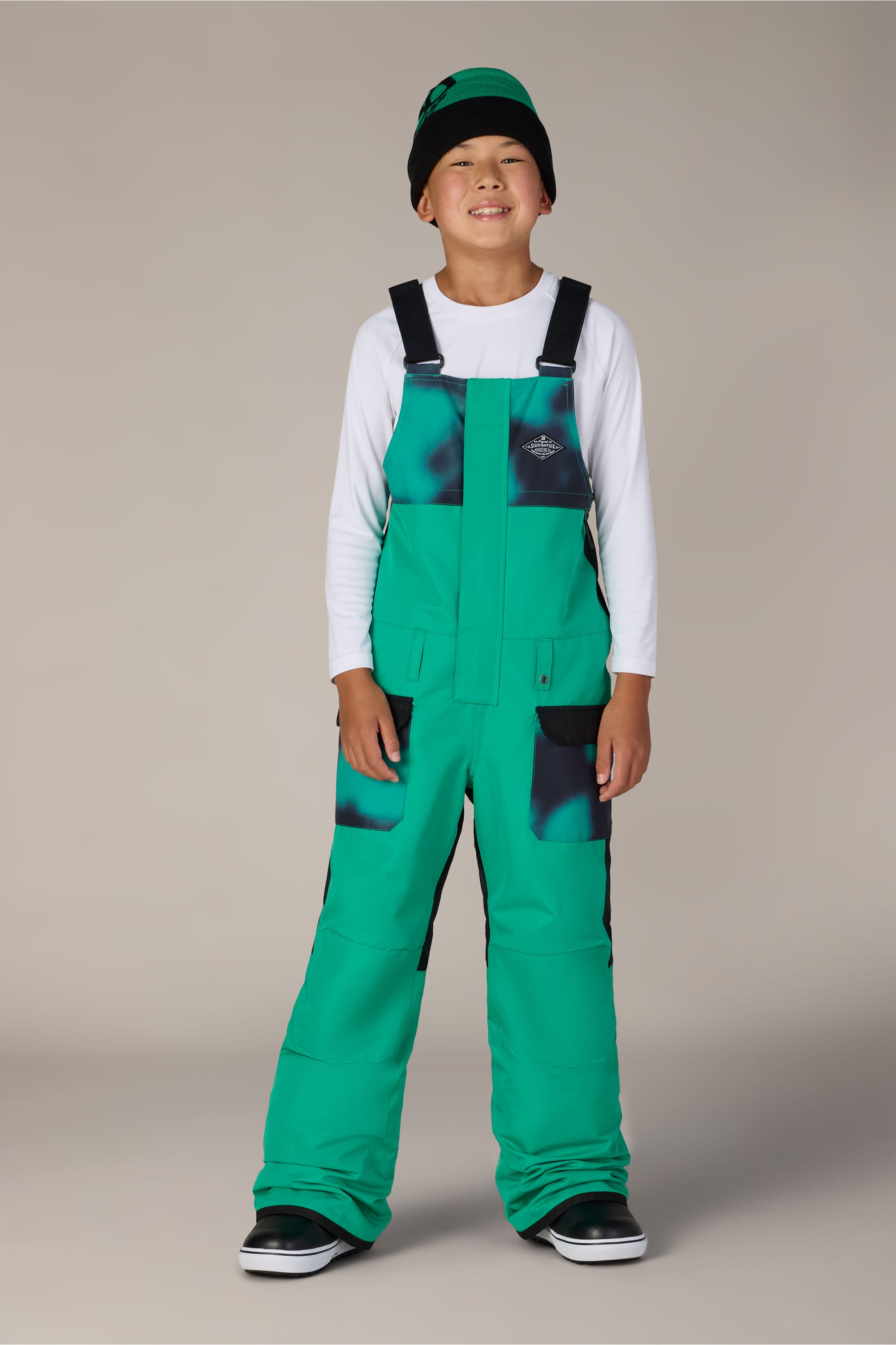 A smiling child wearing the 686 Boys’ Frontier™ Insulated Bib in green with black patches, paired with a green and black beanie and a white long-sleeve shirt, stands against a neutral backdrop.