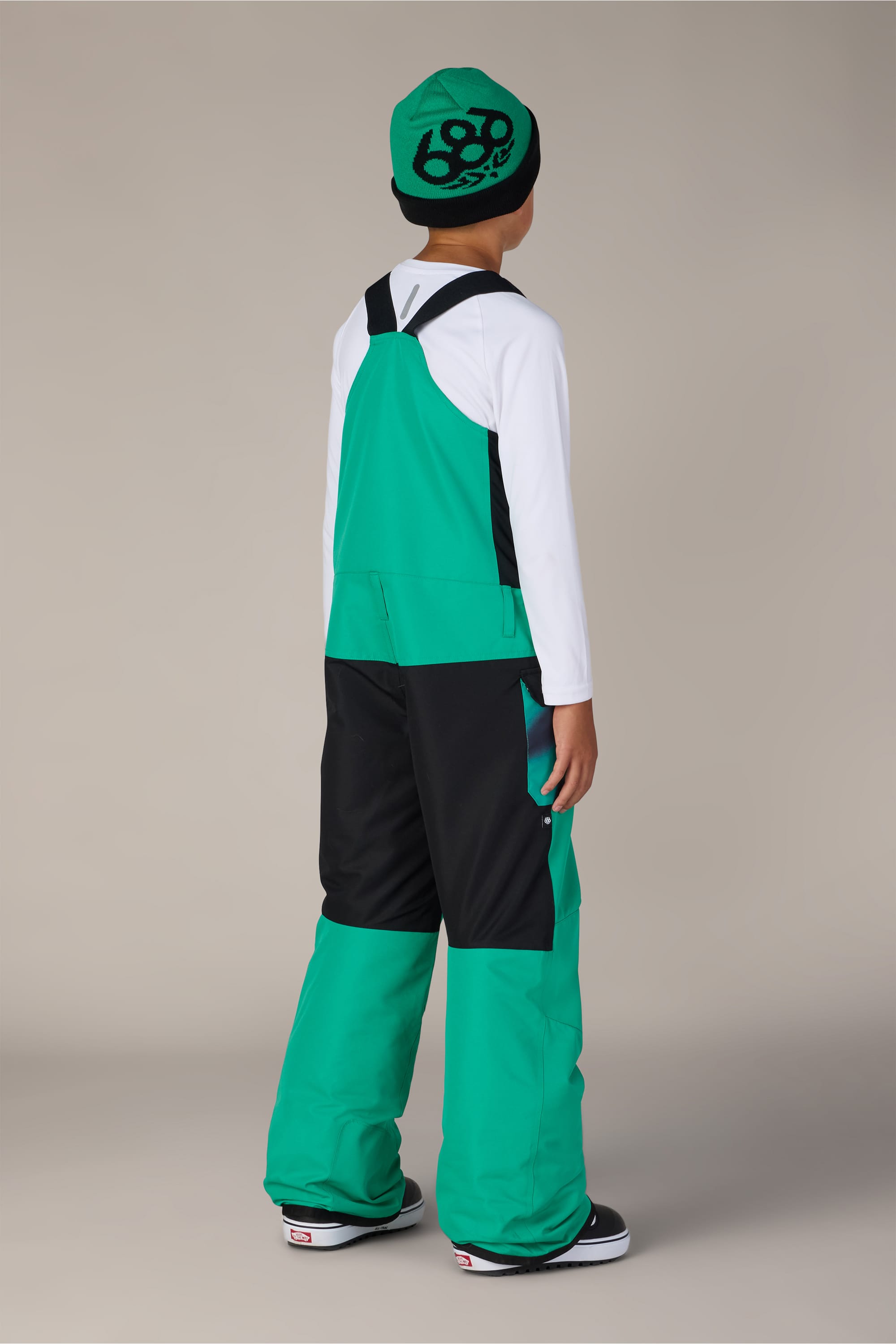 A child wearing the 686 Boys’ Frontier™ Insulated Bib in green and black, a white long-sleeve shirt, a green beanie with a black design, and sneakers stands facing away from the camera.