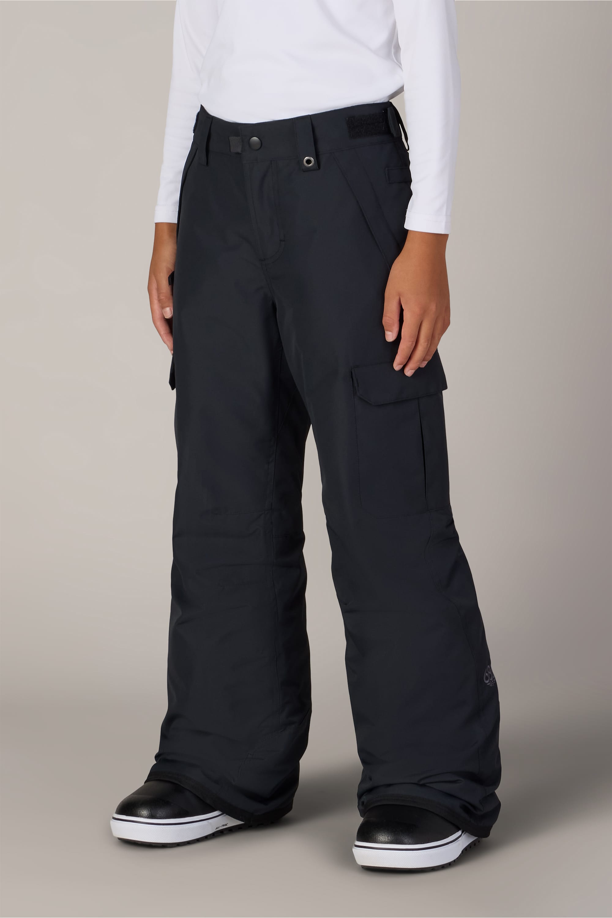 Wearing the 686 Boys Infinity™ Cargo Insulated Pant by 686 in black, a person pairs it with a white long-sleeve shirt and black-and-white shoes while standing against a light gray background.