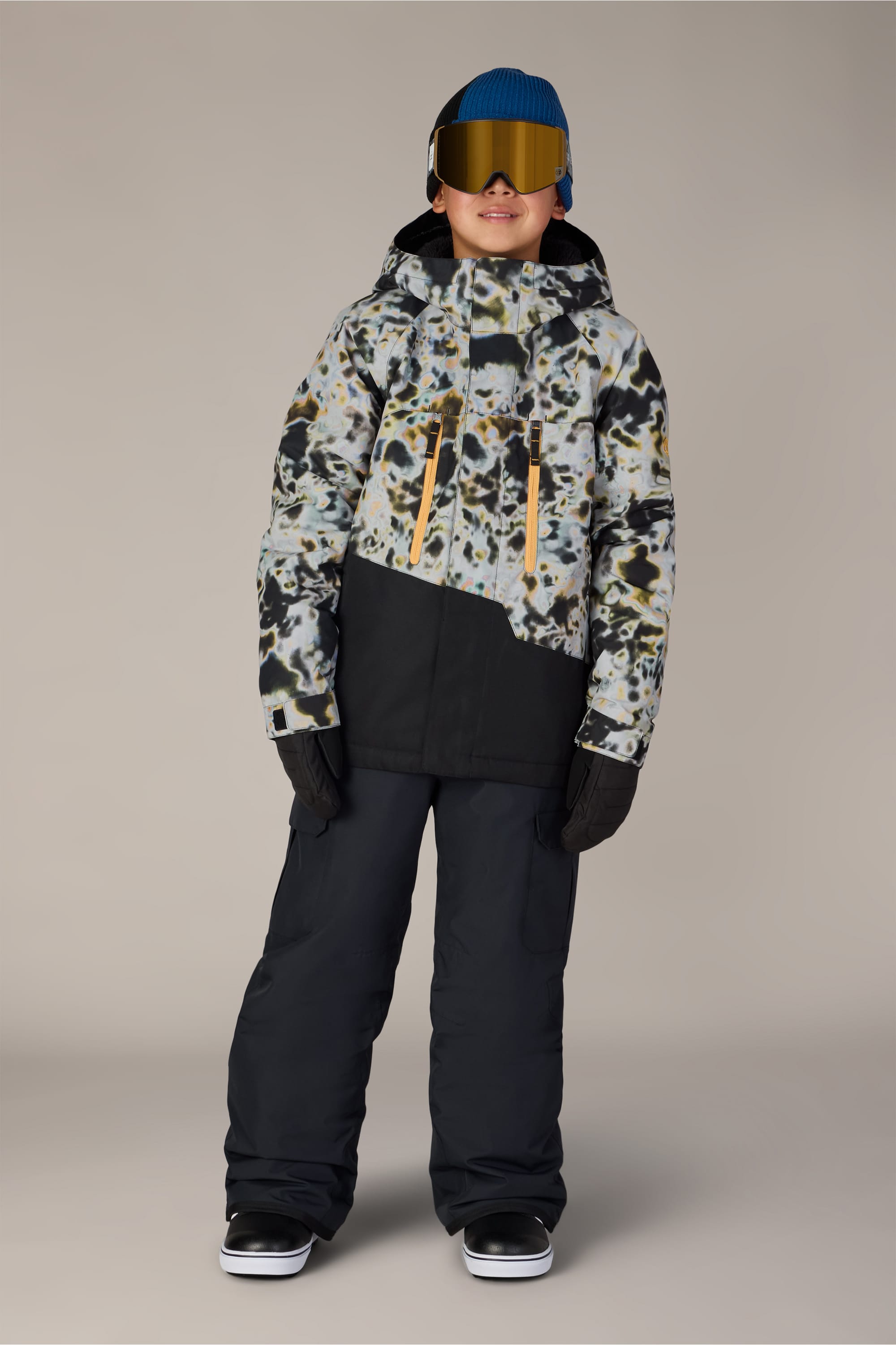A child wearing the 686 Boys Infinity™ Cargo Insulated Pant by 686, along with a patterned winter jacket, blue beanie, ski goggles, and black gloves, stands facing forward against a plain background.