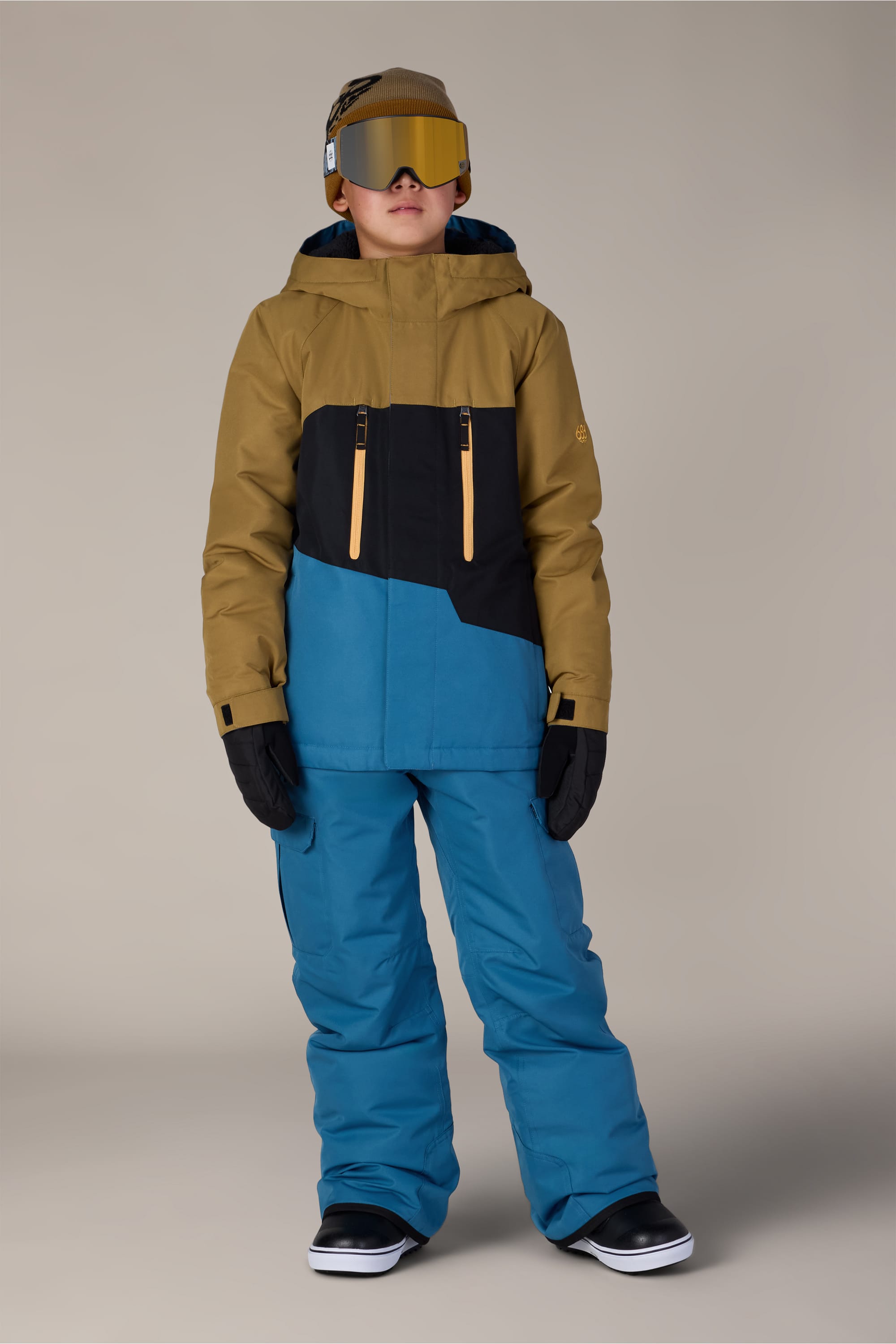 A child wearing a tan, black, and blue winter jacket, 686 Boys Infinity™ Cargo Insulated Pant in blue by 686, black gloves, a tan beanie, ski goggles, and black boots stands facing forward against a plain background.