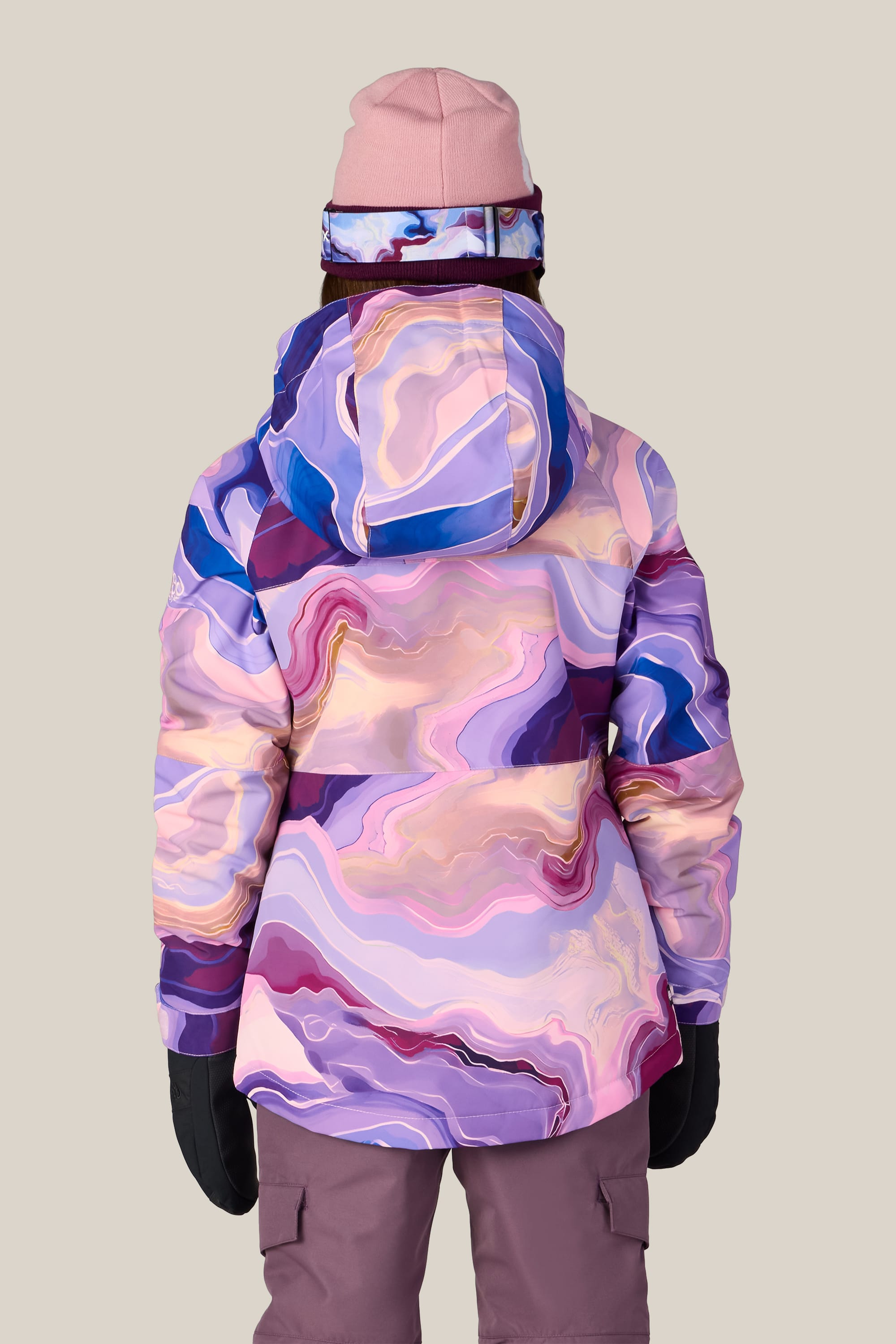 A person is seen from behind, standing against a plain beige background, wearing the 686 Girls’ Athena™ Insulated Jacket by 686—a pink and purple marbled jacket—paired with mauve pants, black gloves, a pink beanie, and ski goggles featuring a matching marbled strap.