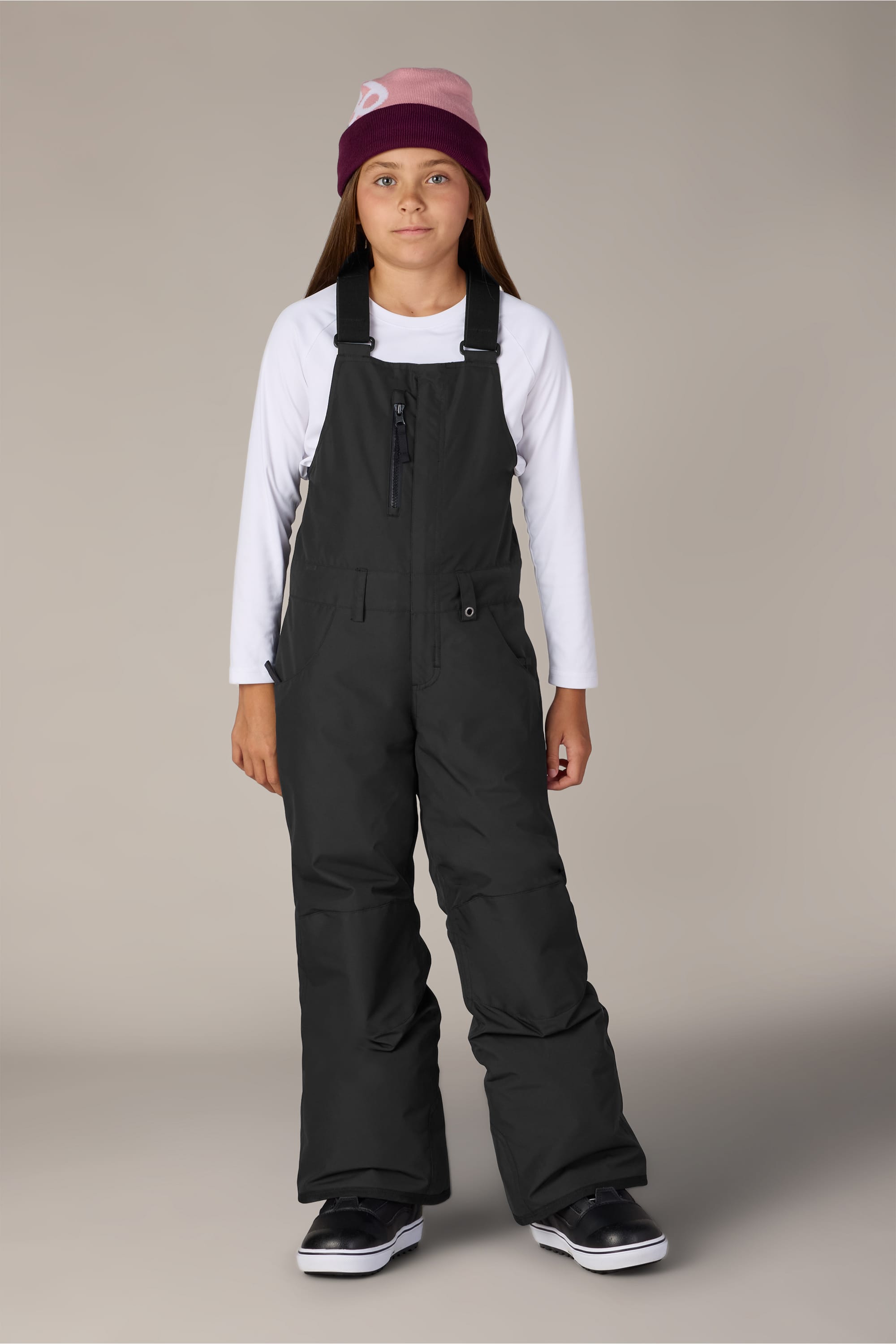 A child wearing the 686 Girls Sierra Insulated Bib by 686 stands facing forward against a plain light gray background, paired with a maroon and pink beanie and a white long-sleeve shirt.