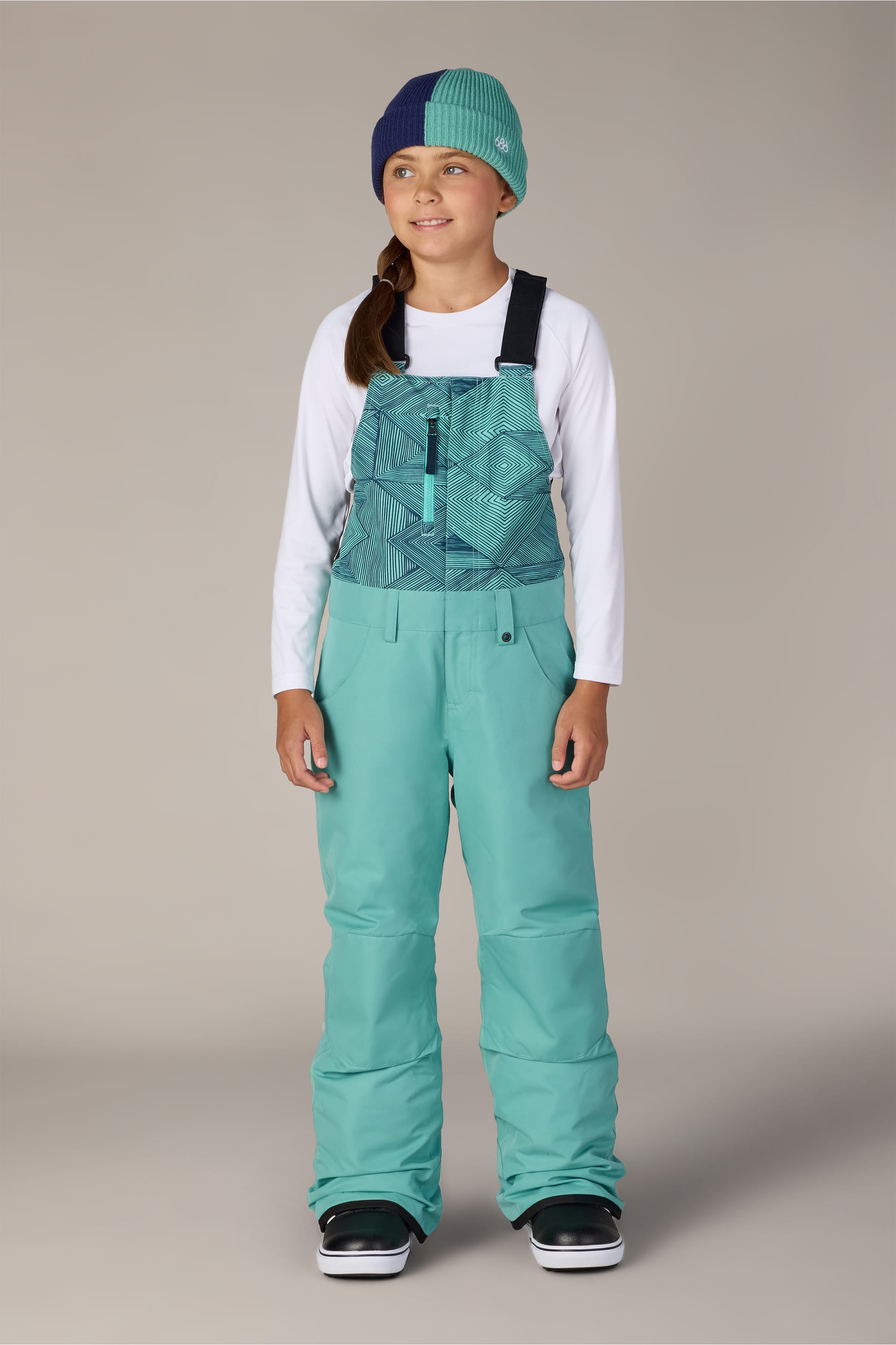 A girl stands facing forward and smiling against a neutral background, wearing the 686 Girls Sierra Insulated Bib snow pants by 686, paired with a white long-sleeve shirt, a blue and green beanie, and black snow boots.