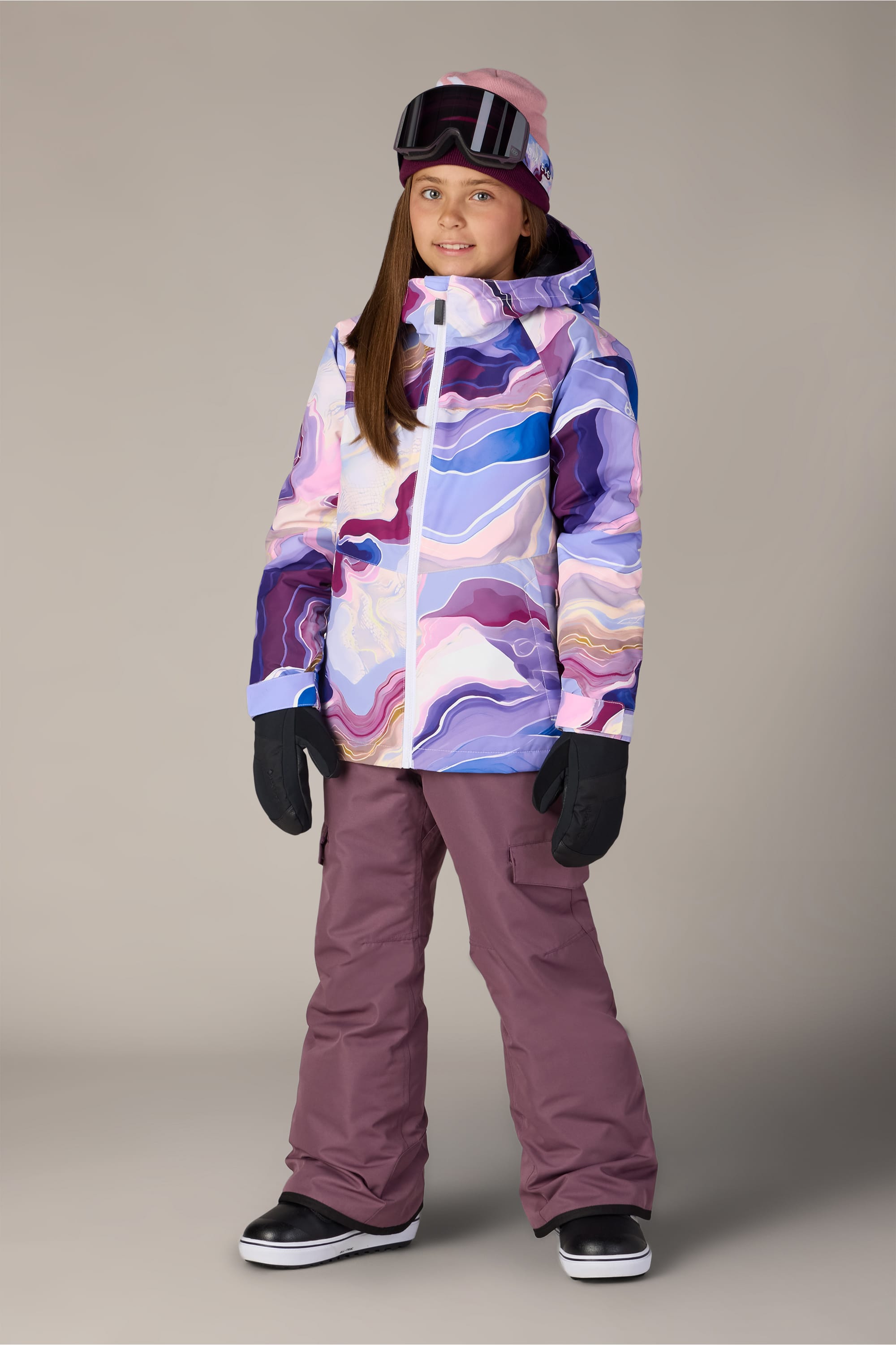 A child wearing a colorful winter jacket, 686 Girls’ Lola Insulated Pant by 686 featuring light insulation and a youth evolution system in maroon, along with black gloves, boots, goggles, and a beanie, stands smiling against a plain background.