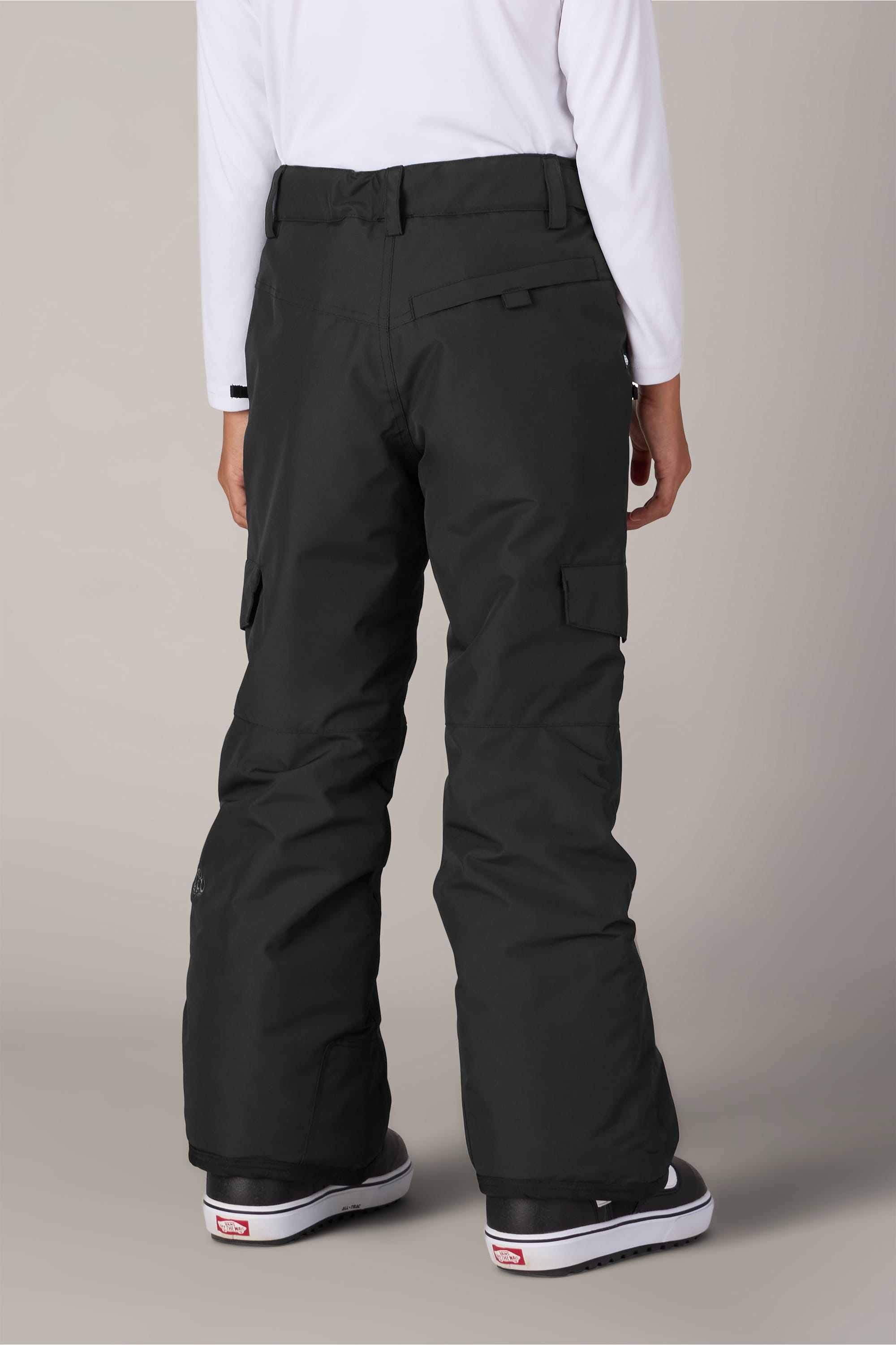 A person is seen from the back, standing against a plain background, wearing the 686 Girls’ Lola Insulated Pant in black for light insulation, paired with a white long-sleeve shirt and black-and-white sneakers.