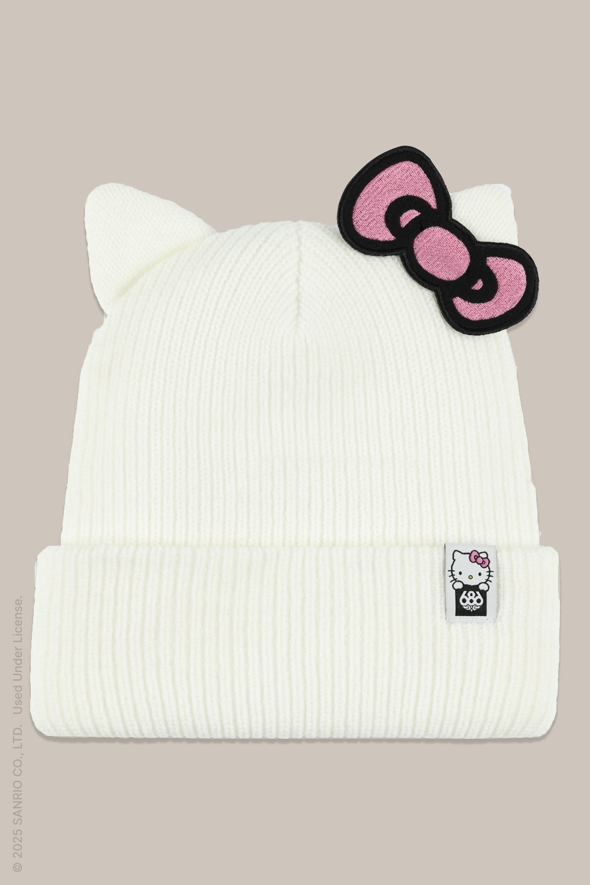 The 686 x Hello Kitty Womens Knit Beanie by 686 is a white knit hat with cat ears, a pink and black bow on one ear, and a Hello Kitty patch on the front cuff, all set against a beige background.