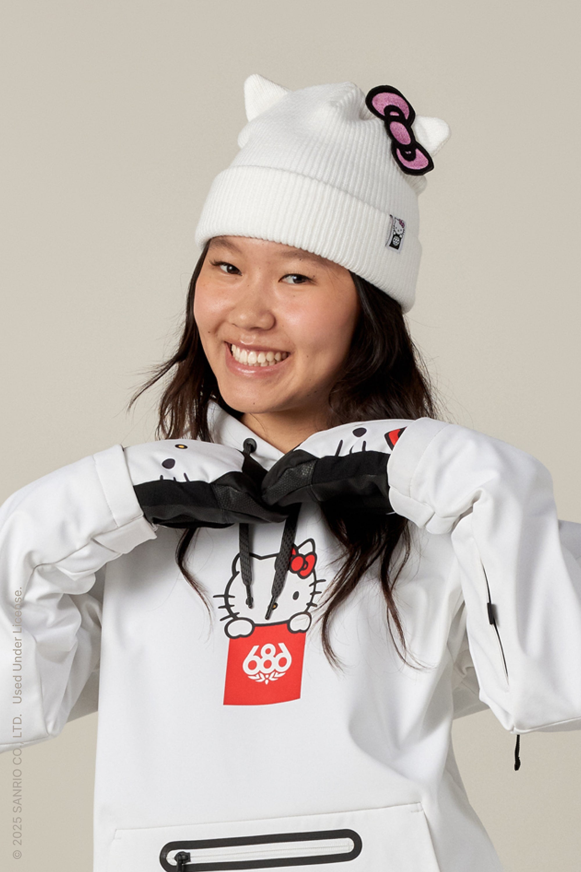A smiling woman combines style and function, wearing the 686 x Hello Kitty Womens Knit Beanie with ears and bow by 686, plus a white hoodie and gloves, posing with her gloved hands under her chin.
