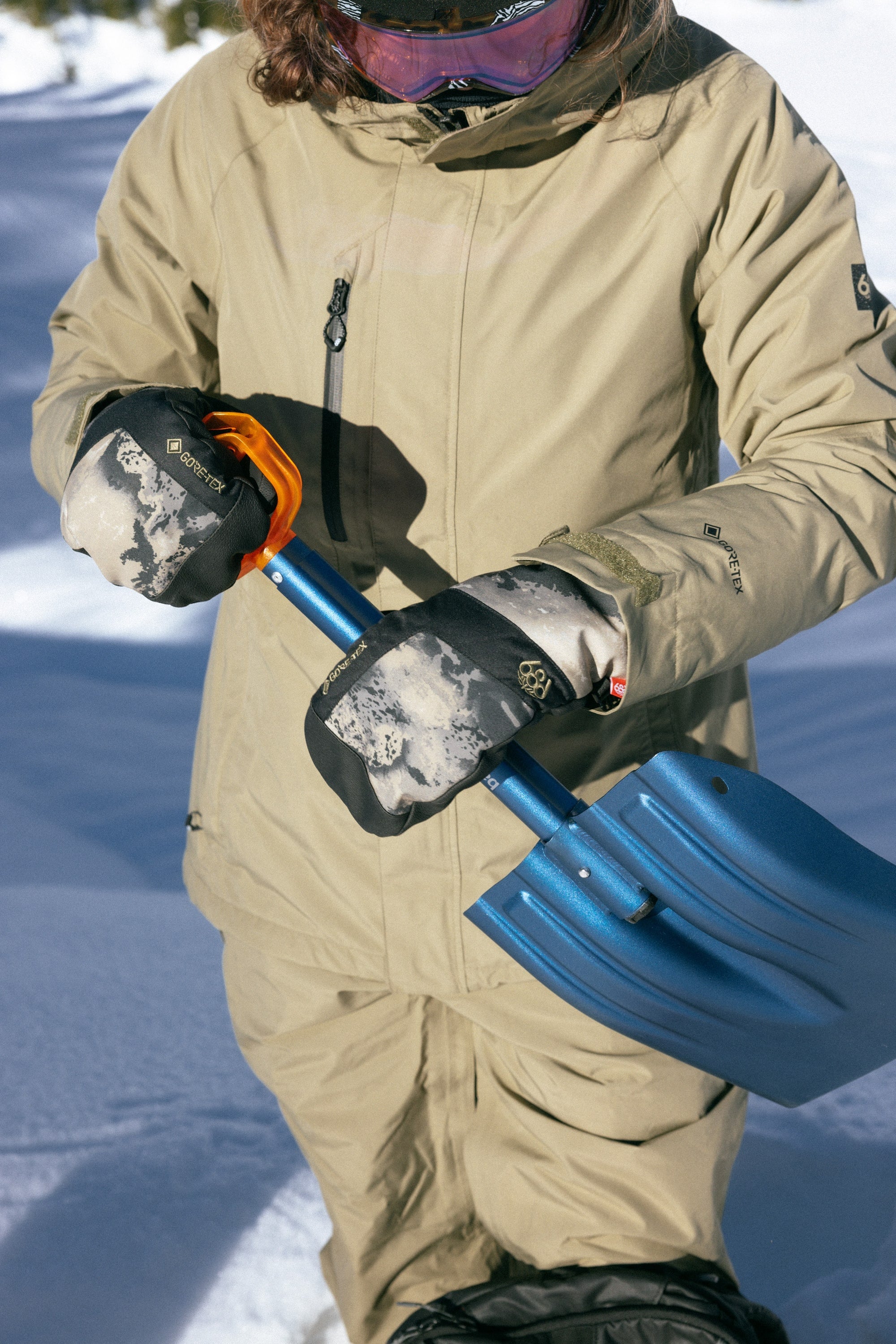 Wearing a tan winter jacket and 686 Mens GORE-TEX Linear Under Cuff Mitt, a person grips a blue snow shovel on snowy ground, ready to tackle the snow with 686’s superior waterproofing. Their face is partially out of frame.