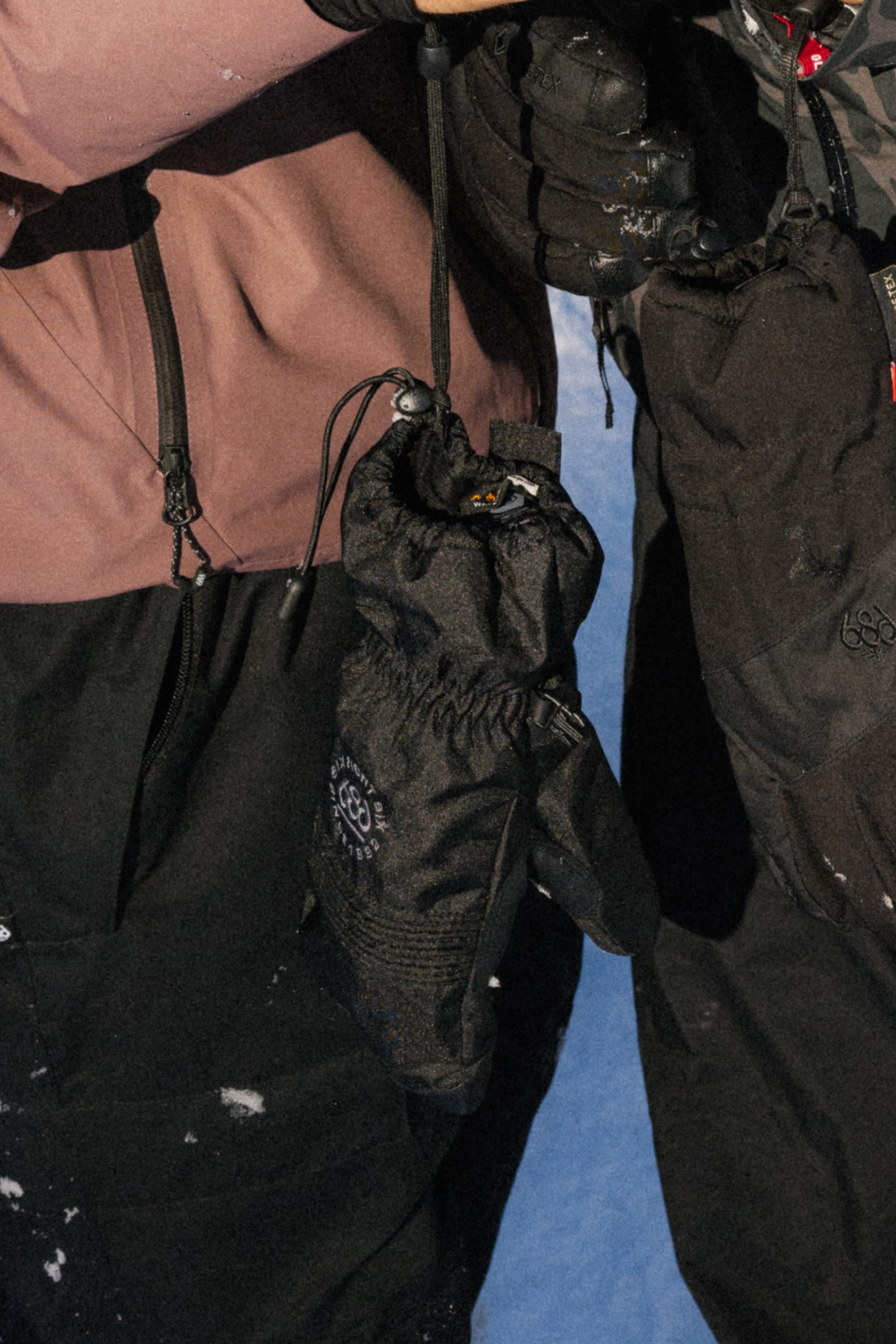 Close-up of two people in winter jackets and snow gloves, holding 686 Lander Mitts by 686, black with drawstrings and 10k waterproofing, against a snowy blue background.