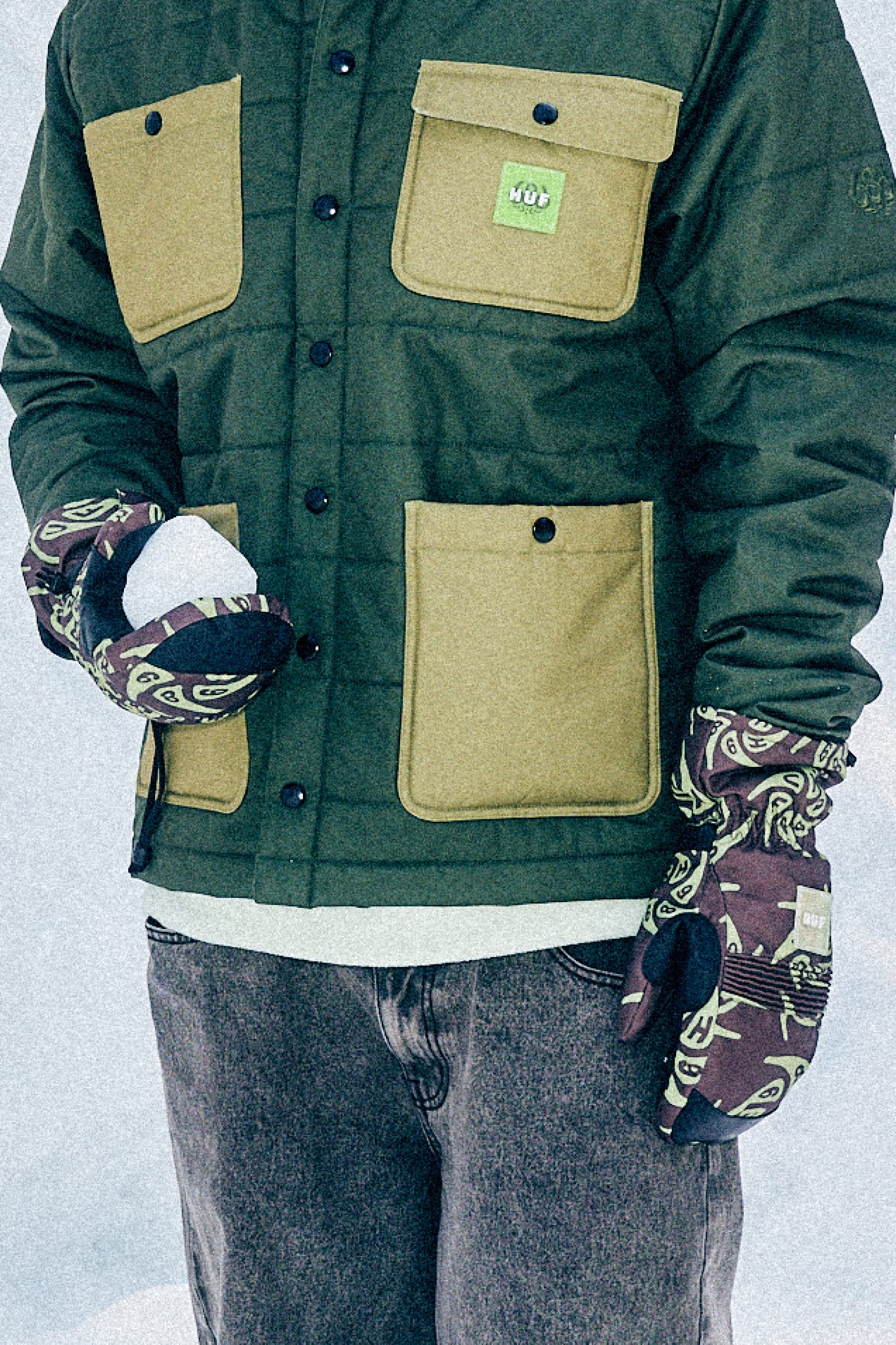 A person in a green and tan lightly insulated jacket, jeans, and 686 Lander Mitts holds a snowball while standing in the snow. Their face is not visible.
