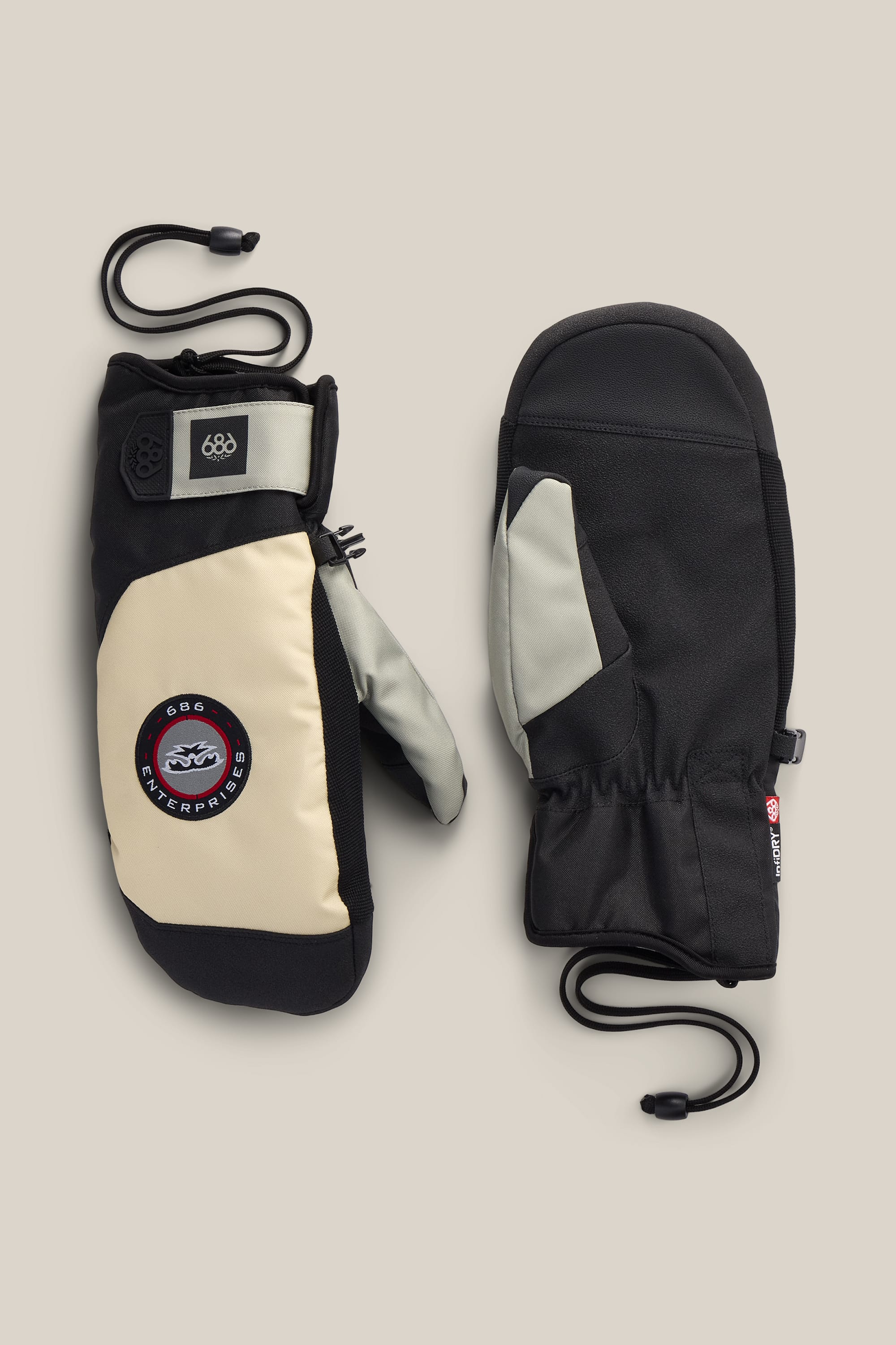 A pair of 686 Men’s Primer Mitts in black and beige, featuring wrist straps and a circular patch labeled “686 Waterproof,” are displayed on a neutral background.