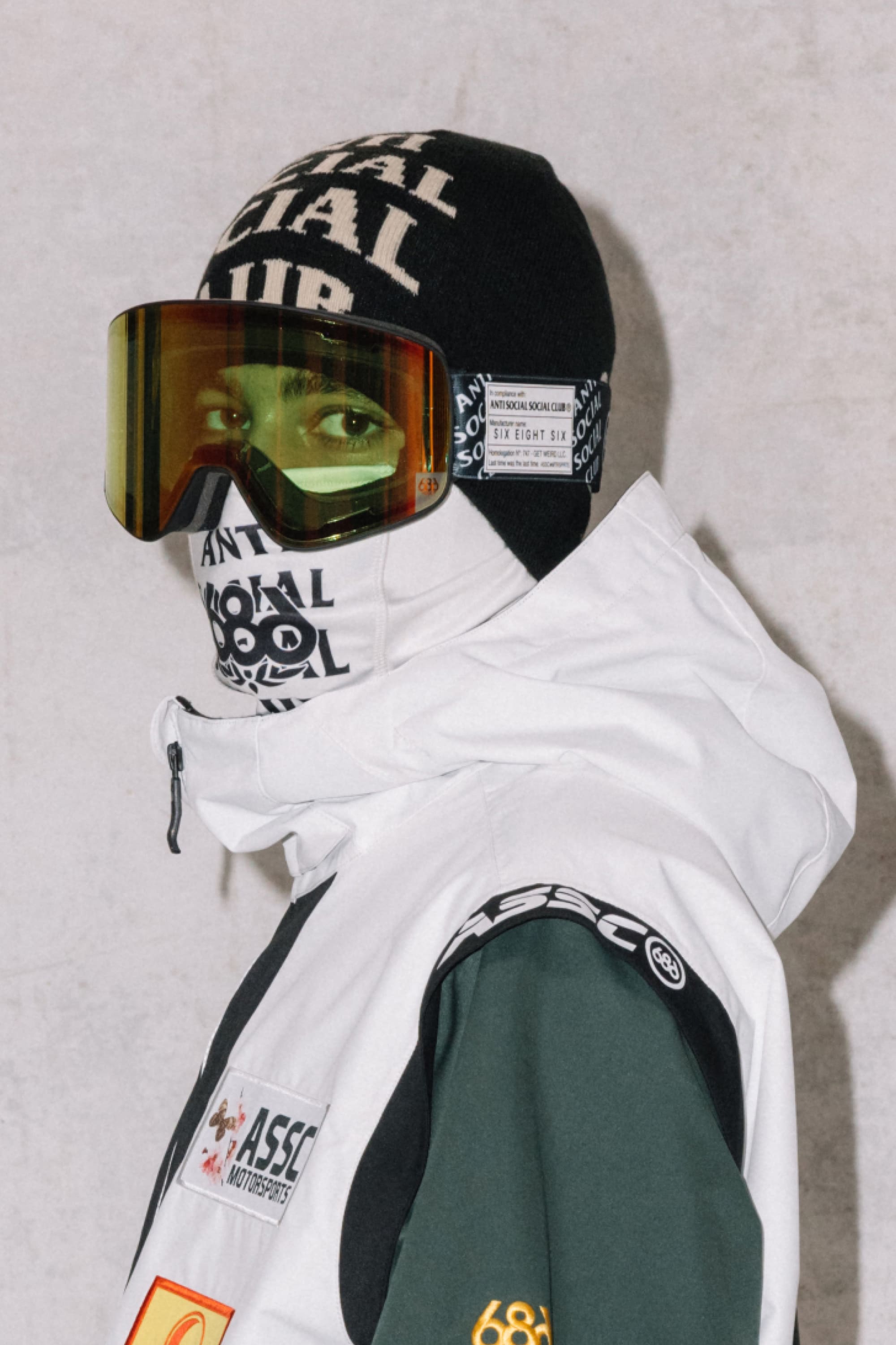 A person wears a black beanie, 686 SMARTY® Magnetic Goggle by 686 in green, and a white jacket with patches. Their face is covered with a black and white ANTI SOCIAL SOCIAL CLUB scarf.
