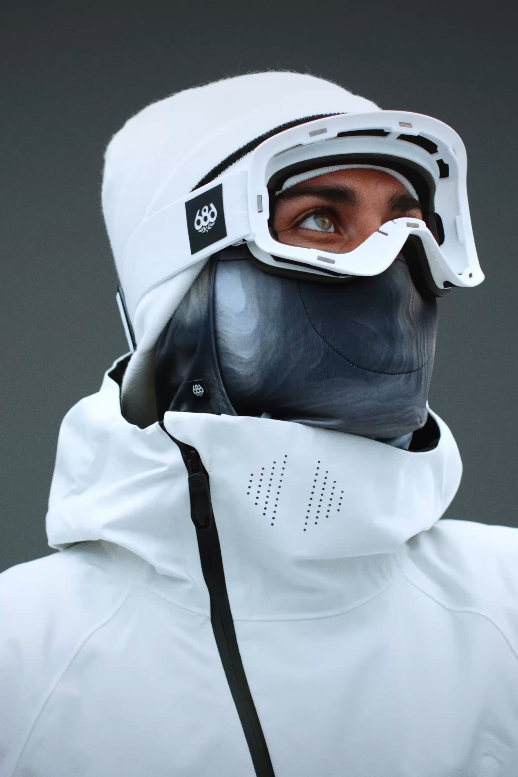 A person in a white jacket, gray face mask, and 686 SMARTY® Magnetic Goggle by 686 looks upward against a dark background, highlighting the winter sports gear.