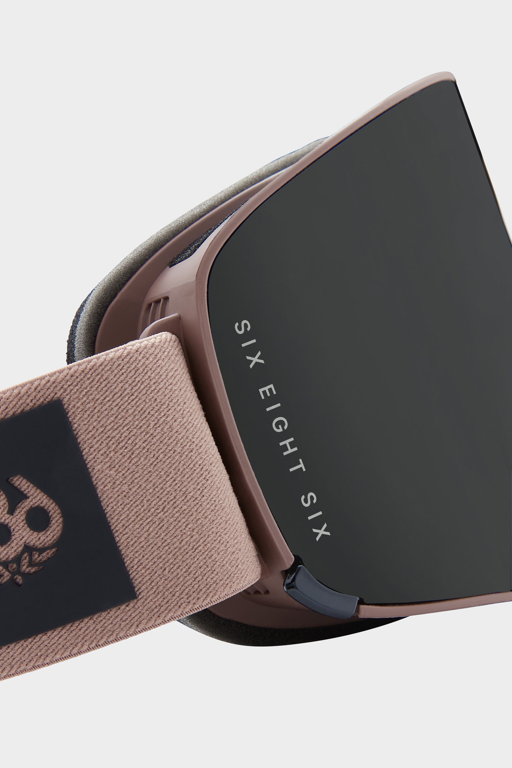 A close-up of the 686 SMARTY Goggle by 686 shows its minimalist design with magnetic lenses, a beige strap, and SIX EIGHT SIX on the black lens. These modern goggles stand out against a light background—ideal for all conditions.