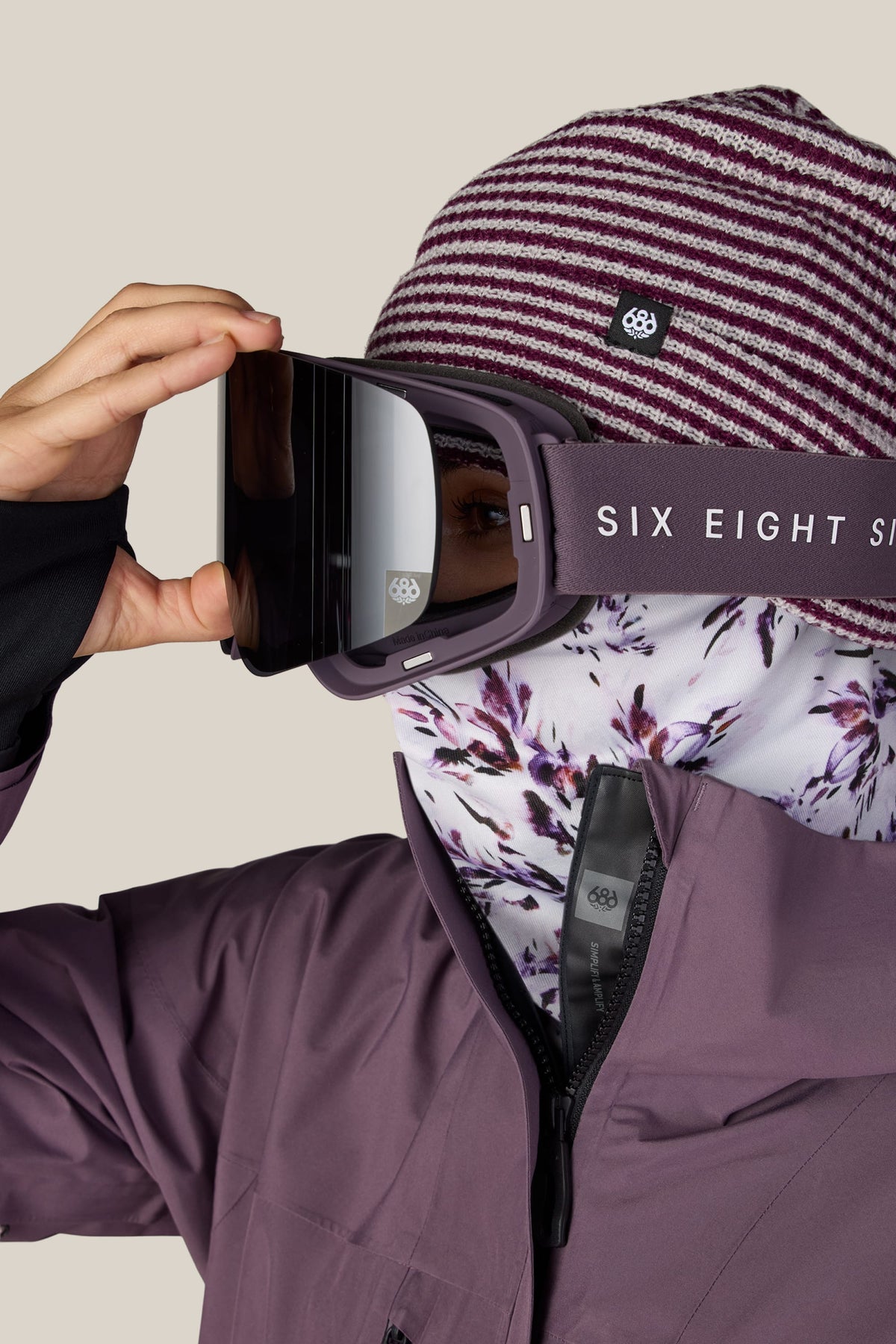 A person in a striped maroon beanie, floral face covering, and purple jacket adjusts large 686 SMARTY® Magnetic Goggles against a light background.