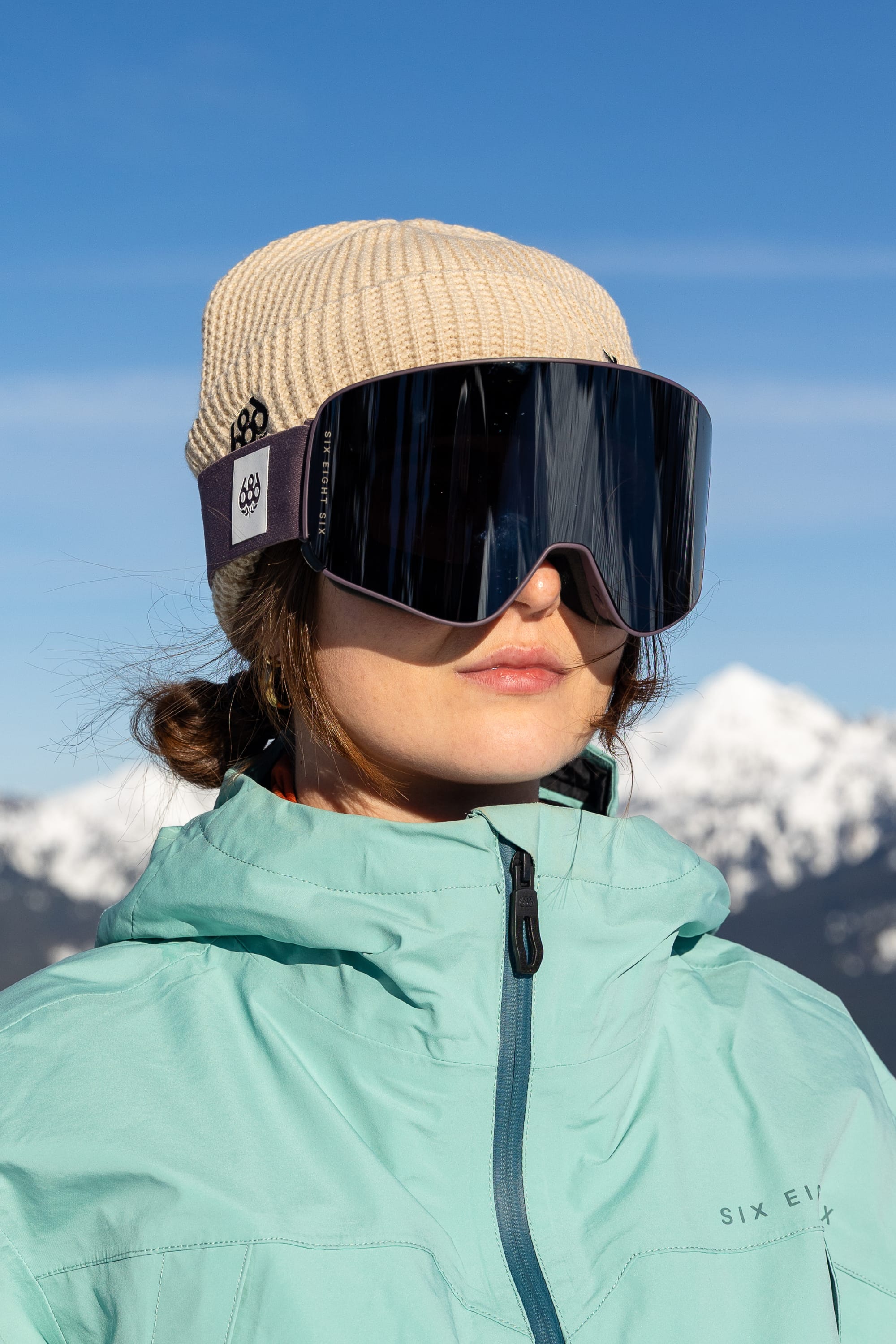 A person stands outdoors against a backdrop of snow-capped mountains and a blue sky, wearing a light blue jacket, tan beanie, and large 686 SMARTY® Magnetic Goggle by 686.