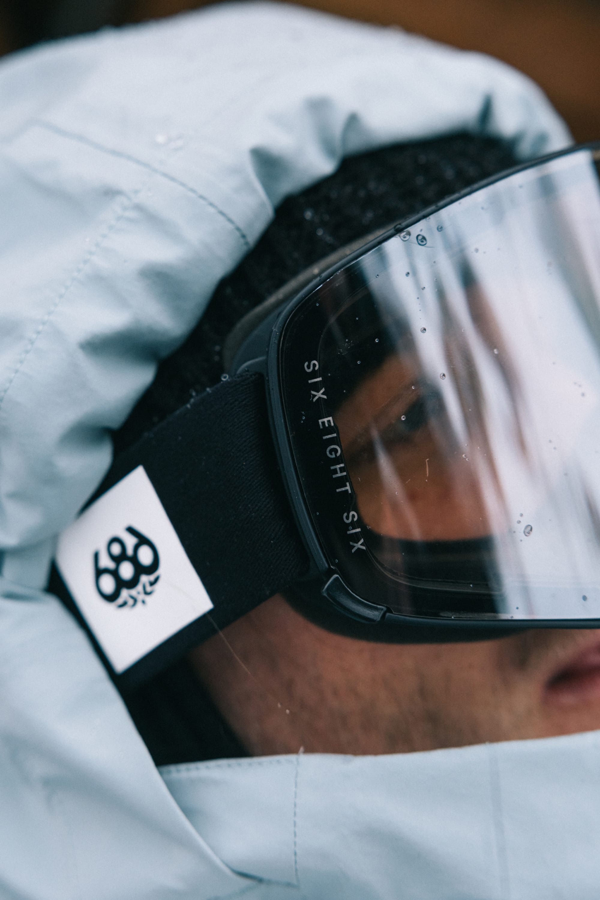 A close-up of a person wearing a blue hooded jacket, black 686 SMARTY® Magnetic Goggle with SIX EIGHT SIX on the strap, and a beanie. The white 686 logo patch is visible on the goggle strap.