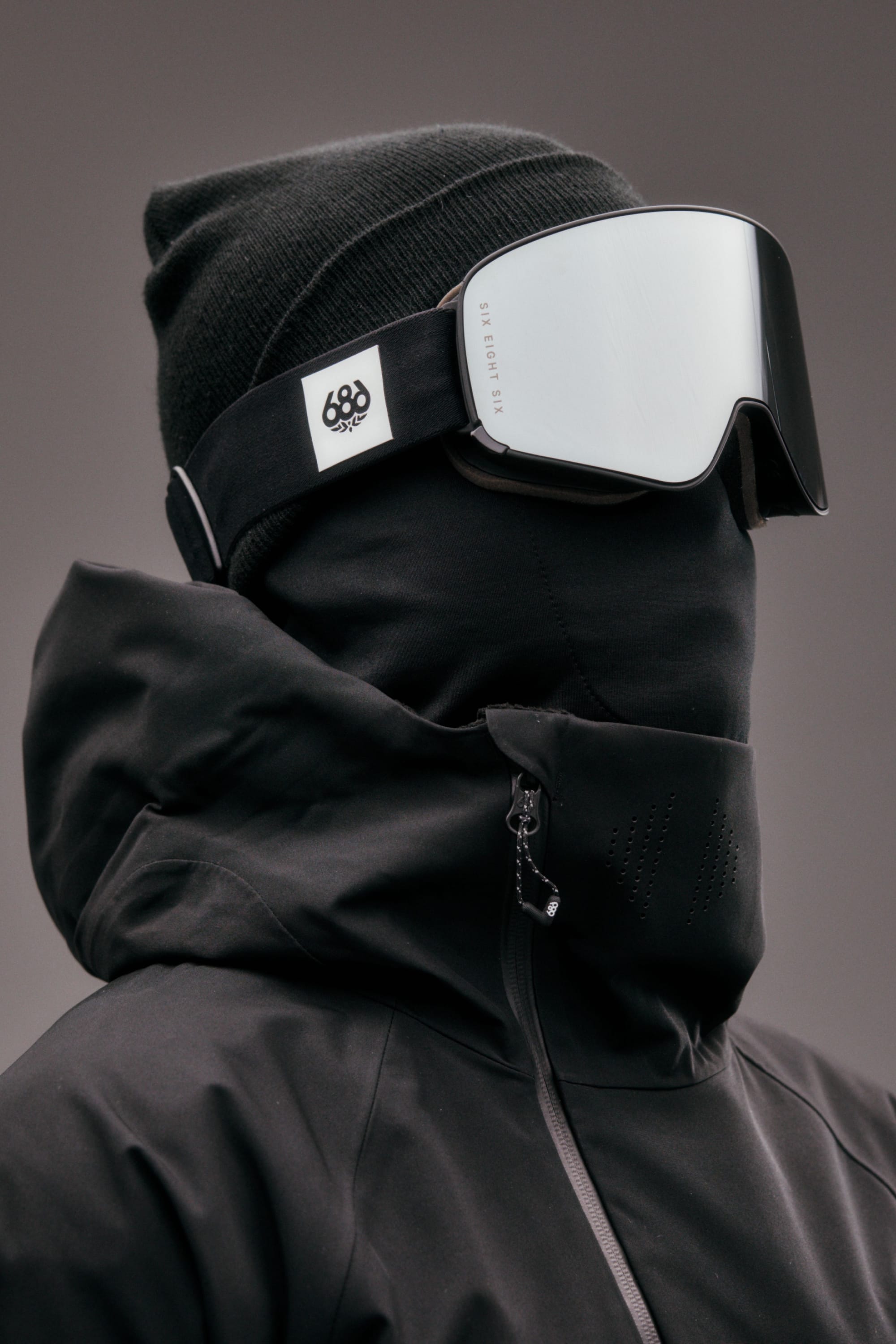 Wearing a black beanie, 686 SMARTY Goggle by 686 with magnetic lenses, and a black face covering under a black jacket, only their nose visible. The plain neutral background suits any condition.