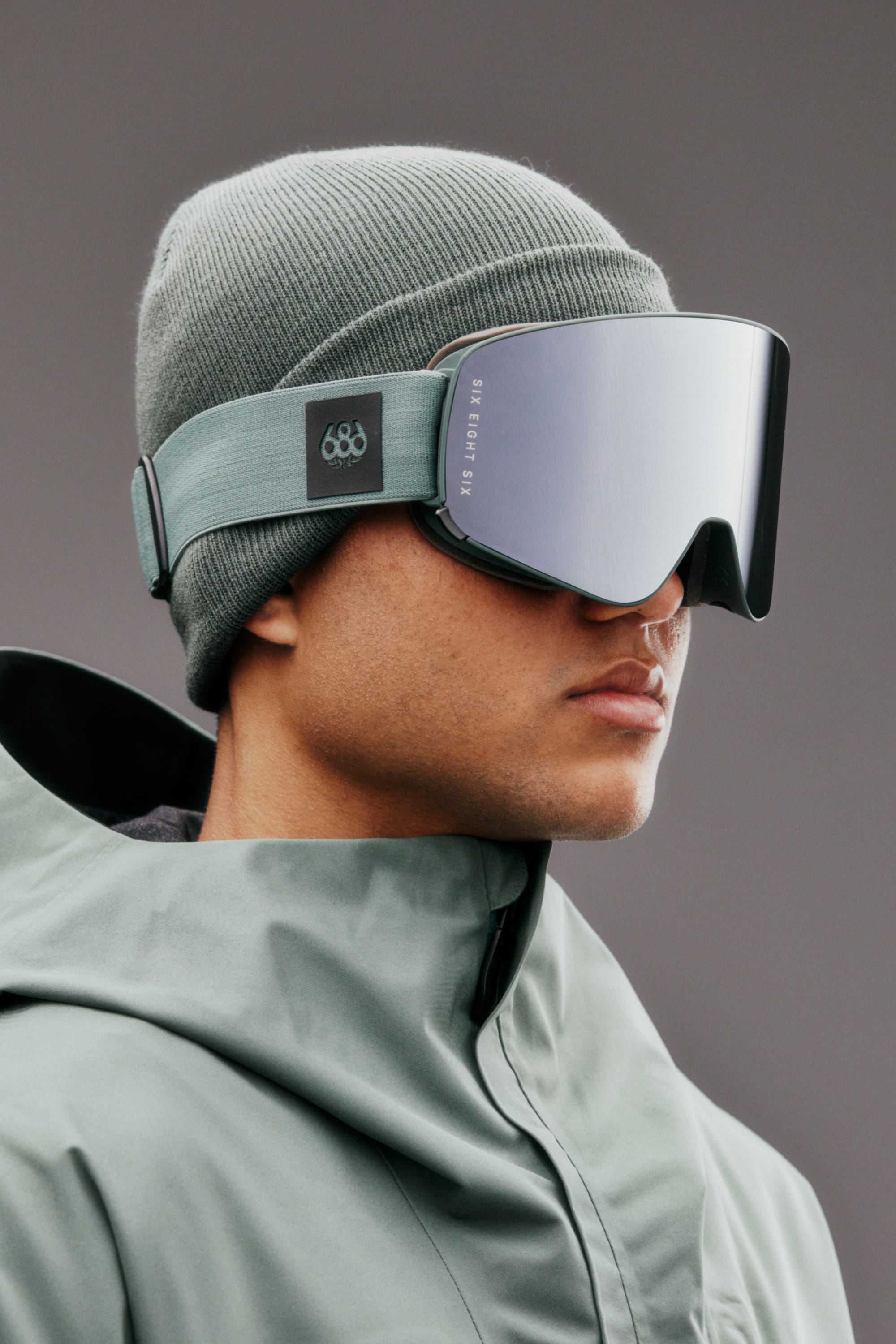 Wearing a green beanie, a 686 SMARTY Goggle by 686 with magnetic lenses, and a matching green jacket, this person poses against a gray background—ready for any conditions.