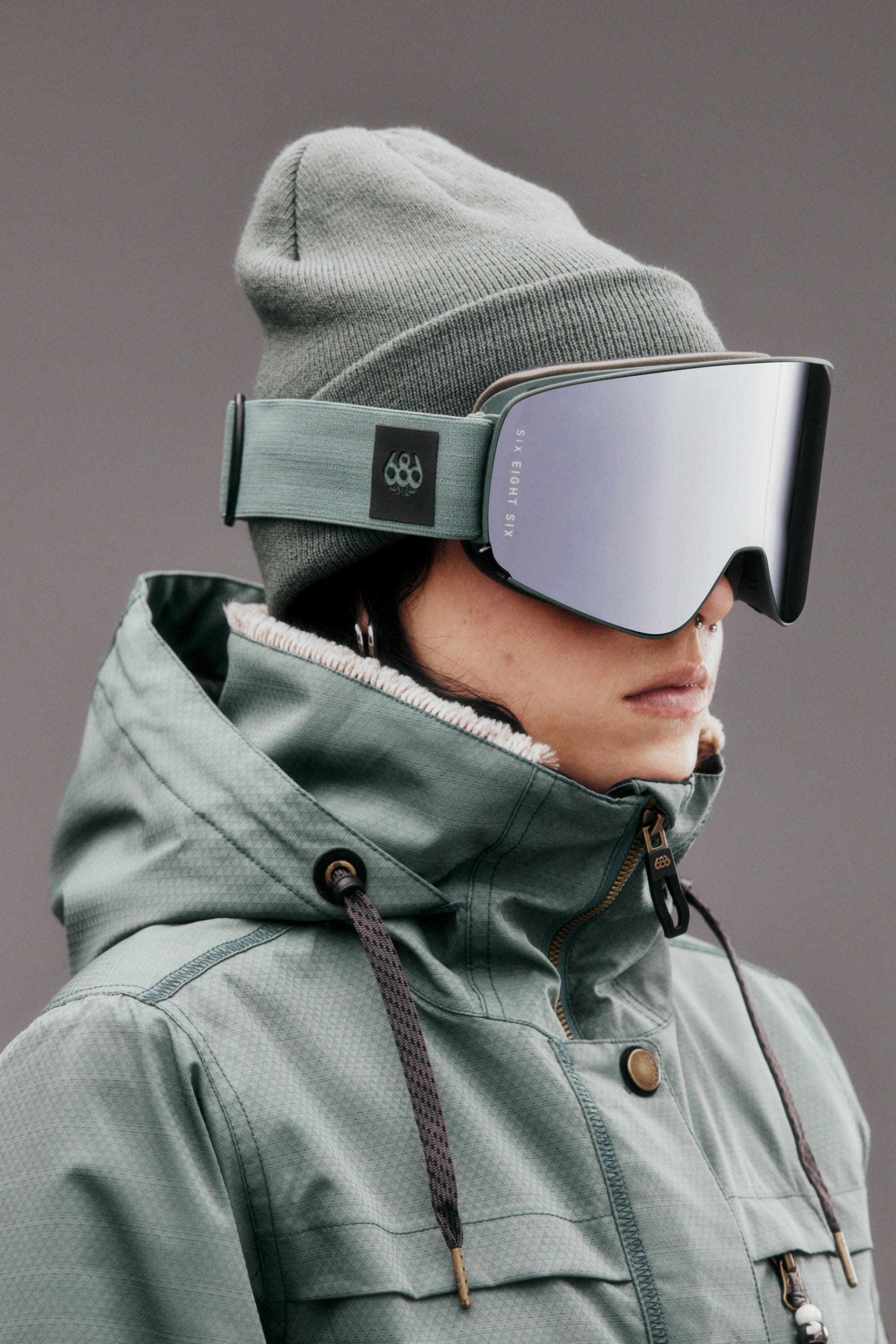 Wearing a green beanie and jacket, the person sports 686 SMARTY Goggle by 686. The large, reflective, minimalist goggles with magnetic lenses stand out against the plain gray background as the person faces sideways.