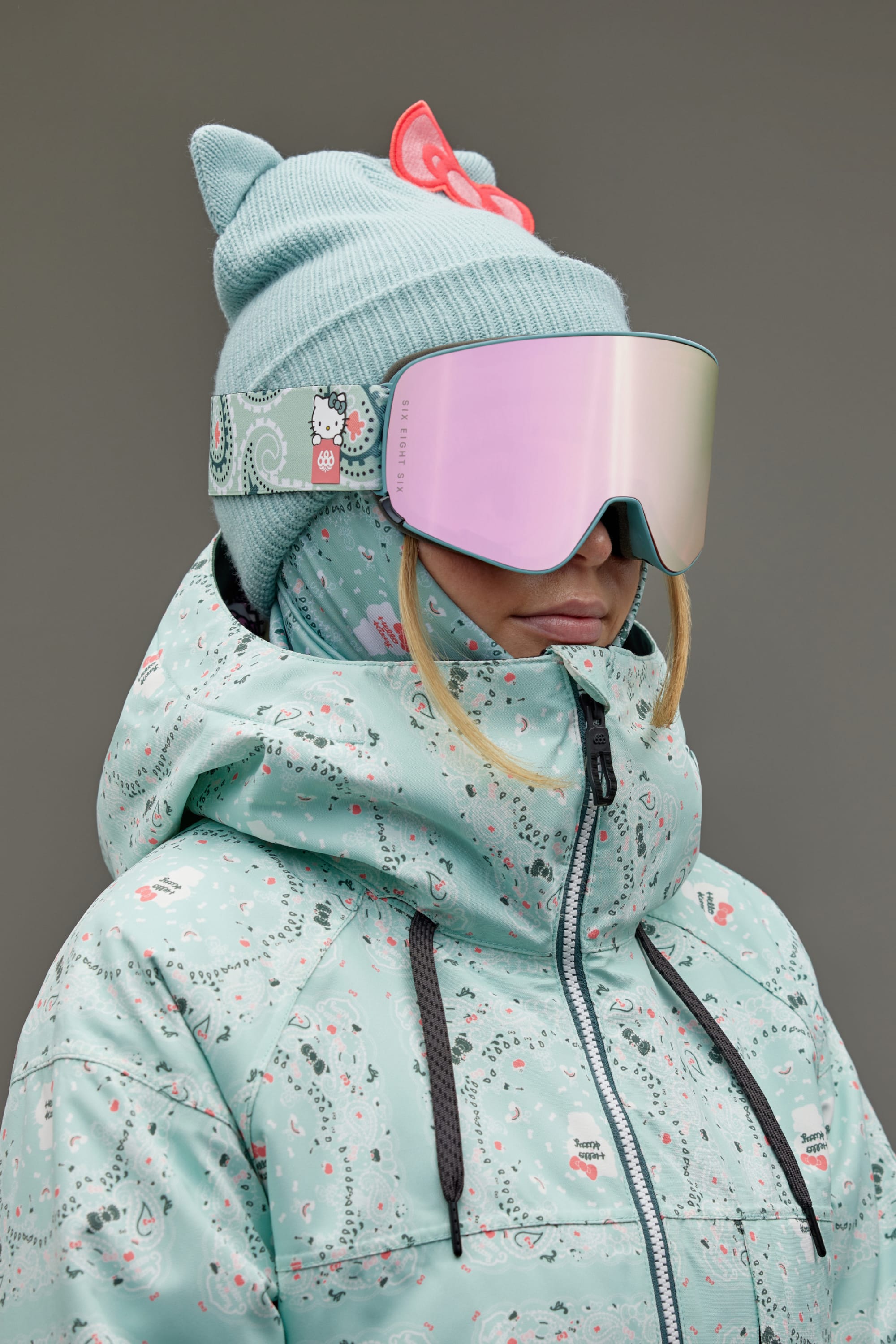 A woman in a pastel blue Hello Kitty ski jacket, 686 SMARTY Goggle by 686 with magnetic lenses for all conditions, and a blue knit hat with cat ears and a red bow stands against a plain gray background.