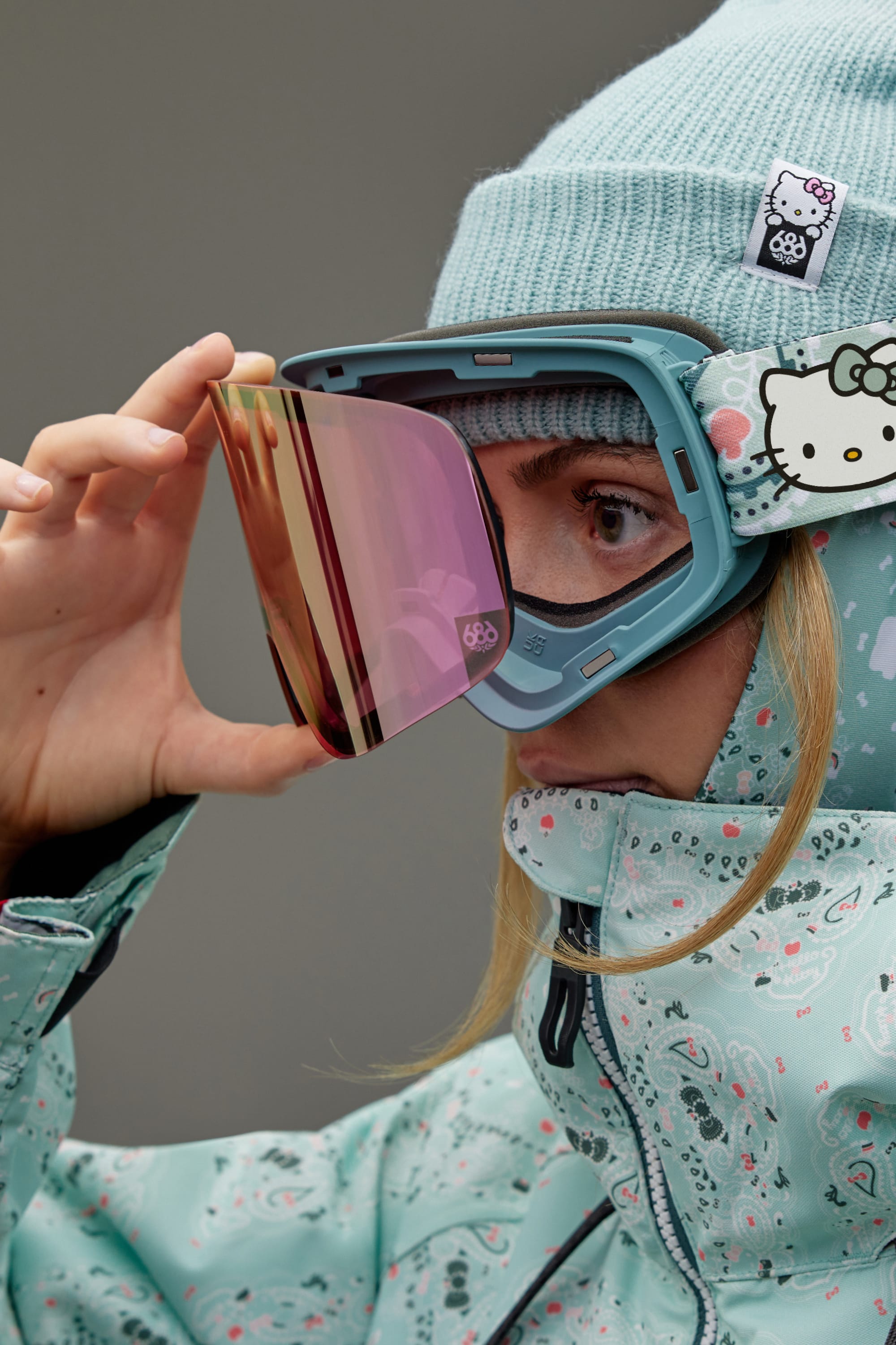 A person in a blue patterned jacket and beanie adjusts the pink magnetic lenses of their 686 SMARTY Goggle by 686, featuring a Hello Kitty design on the strap.