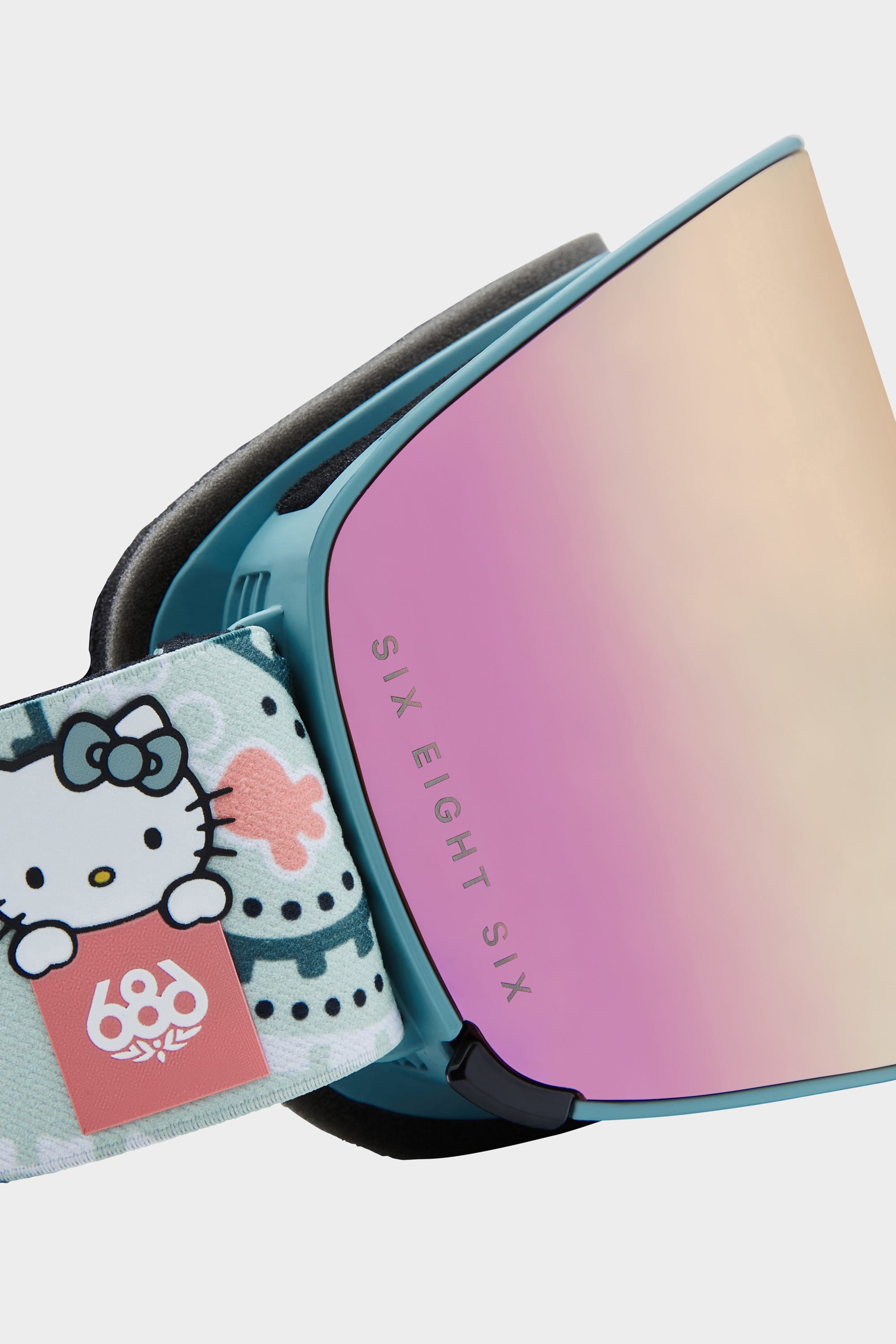 Close-up of teal 686 SMARTY Goggle with pink reflective magnetic lens. The strap shows a Hello Kitty and sea creature design, 686 logo on pastel. SIX EIGHT SIX text on the frame; perfect for all conditions.