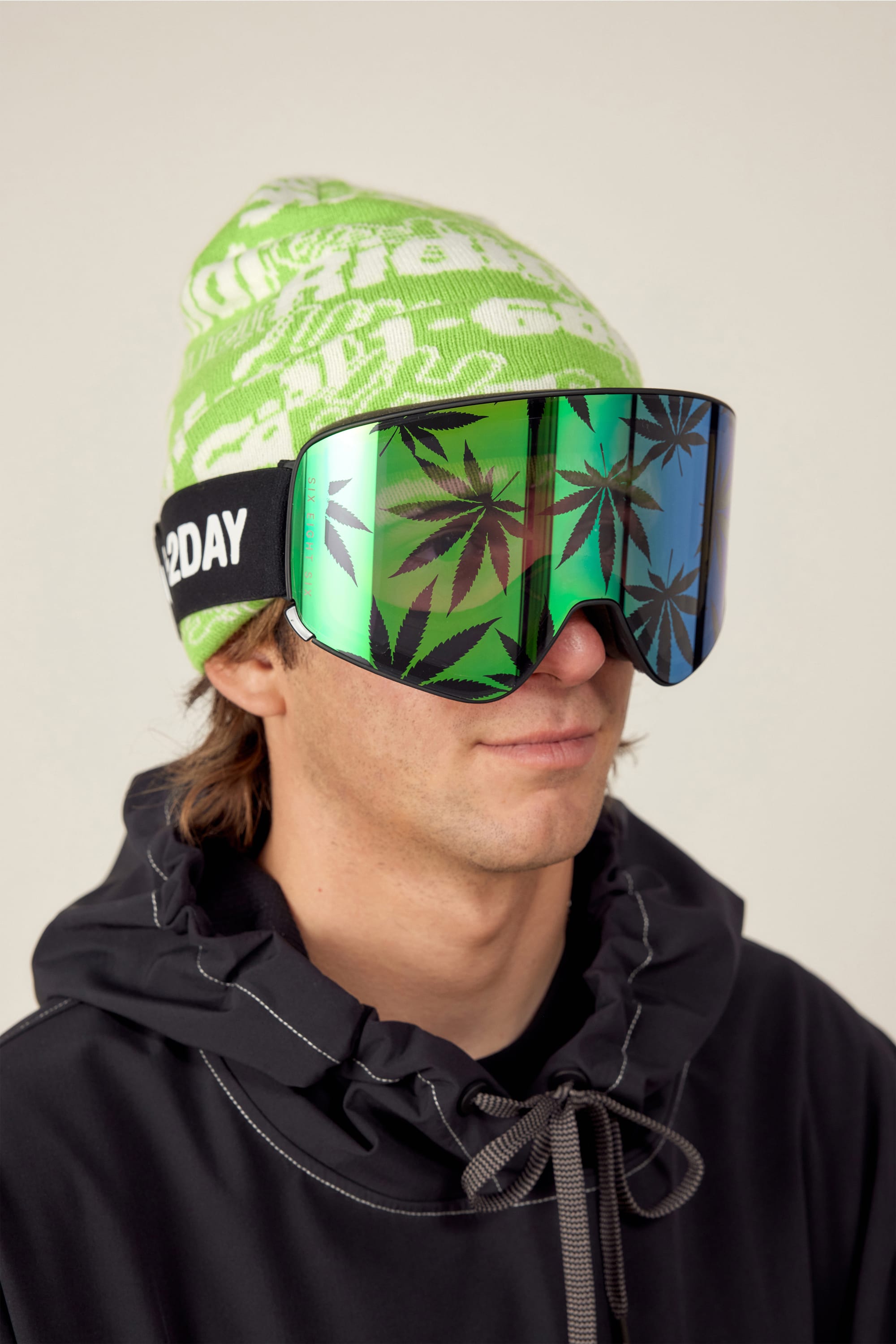 A person wears the 686 SMARTY Goggle by 686 with large reflective lenses featuring black marijuana leaf prints, paired with a neon green patterned beanie and black jacket. The plain, neutral background suits all conditions.